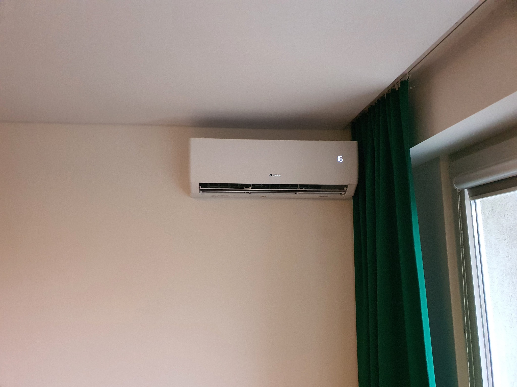 Biała klimatyzacja ścienna z wyświetlaczem temperatury 16 stopni Celsjusza, zamontowana na beżowej ścianie w pokoju z zielonymi zasłonami i oknem.