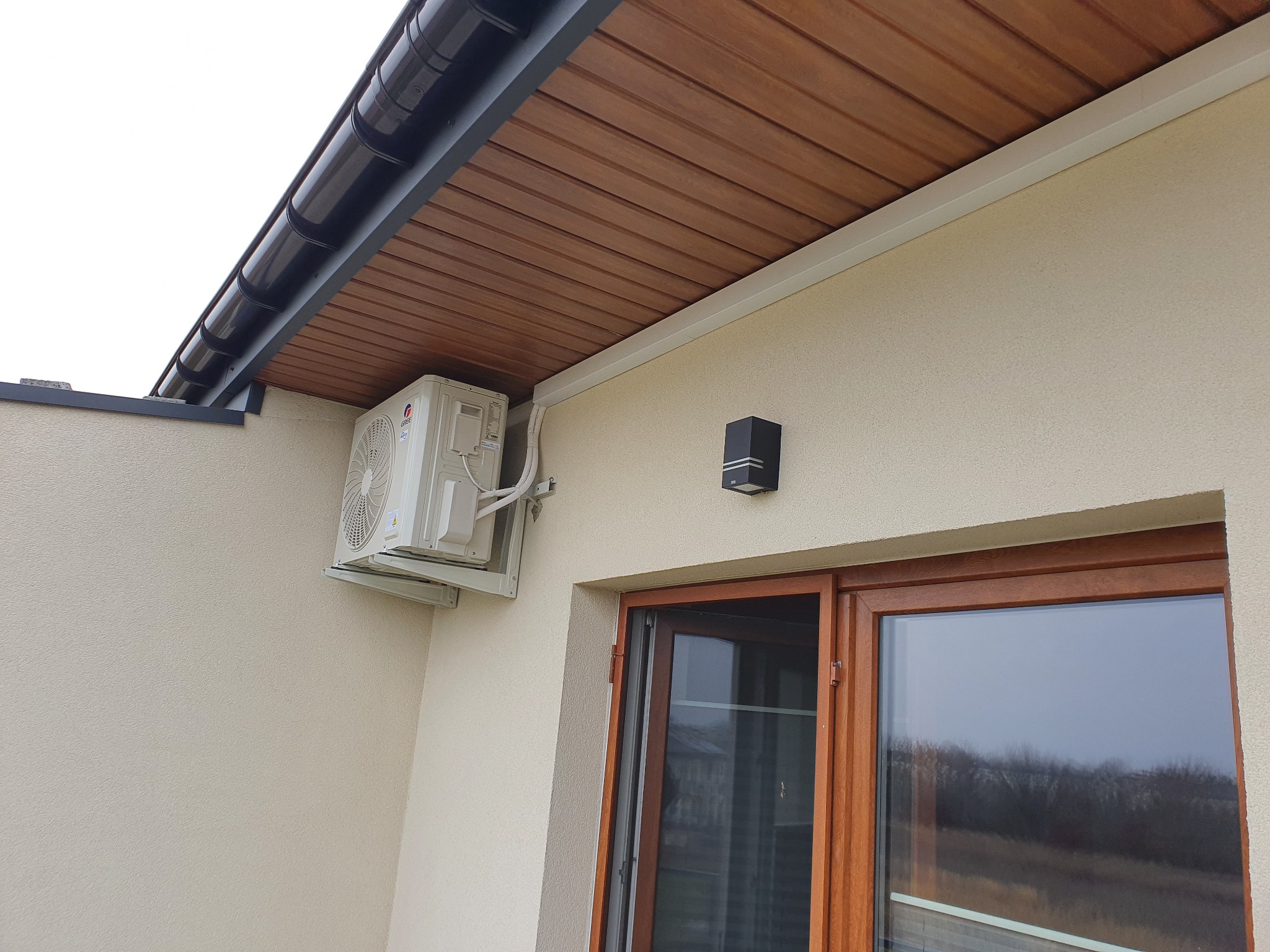 Zewnętrzna jednostka klimatyzatora zamontowana na elewacji domu, widoczne okno z brązowymi ramami i czarny kinkiet.