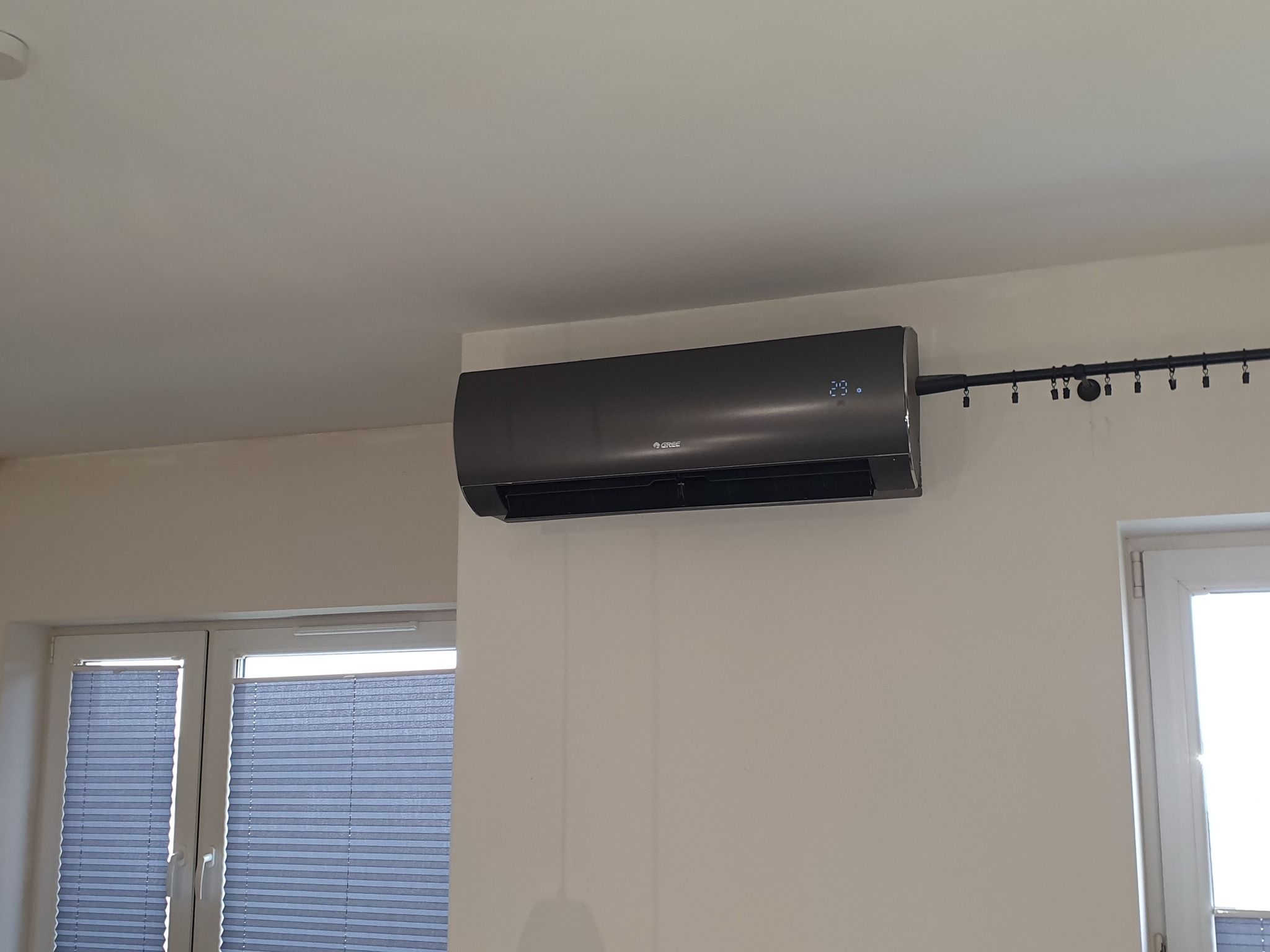 Czarny klimatyzator Gree zamontowany na białej ścianie w pomieszczeniu z oknem z żaluzjami plisowanymi, widoczny czarny karnisz z klipsami.