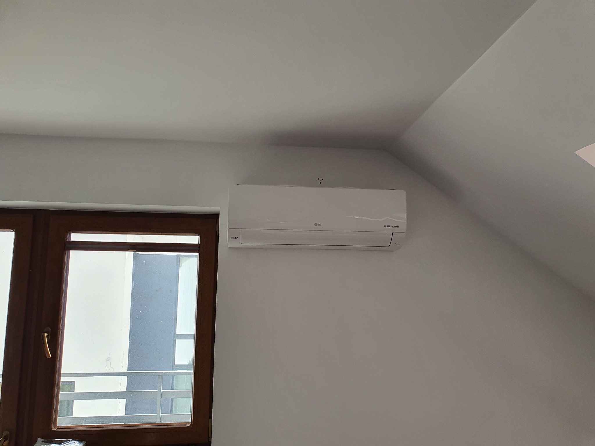 Biała jednostka klimatyzacji LG Dual Inverter zamontowana na białej ścianie pod skosem, obok drewnianego okna z widokiem na balkon.