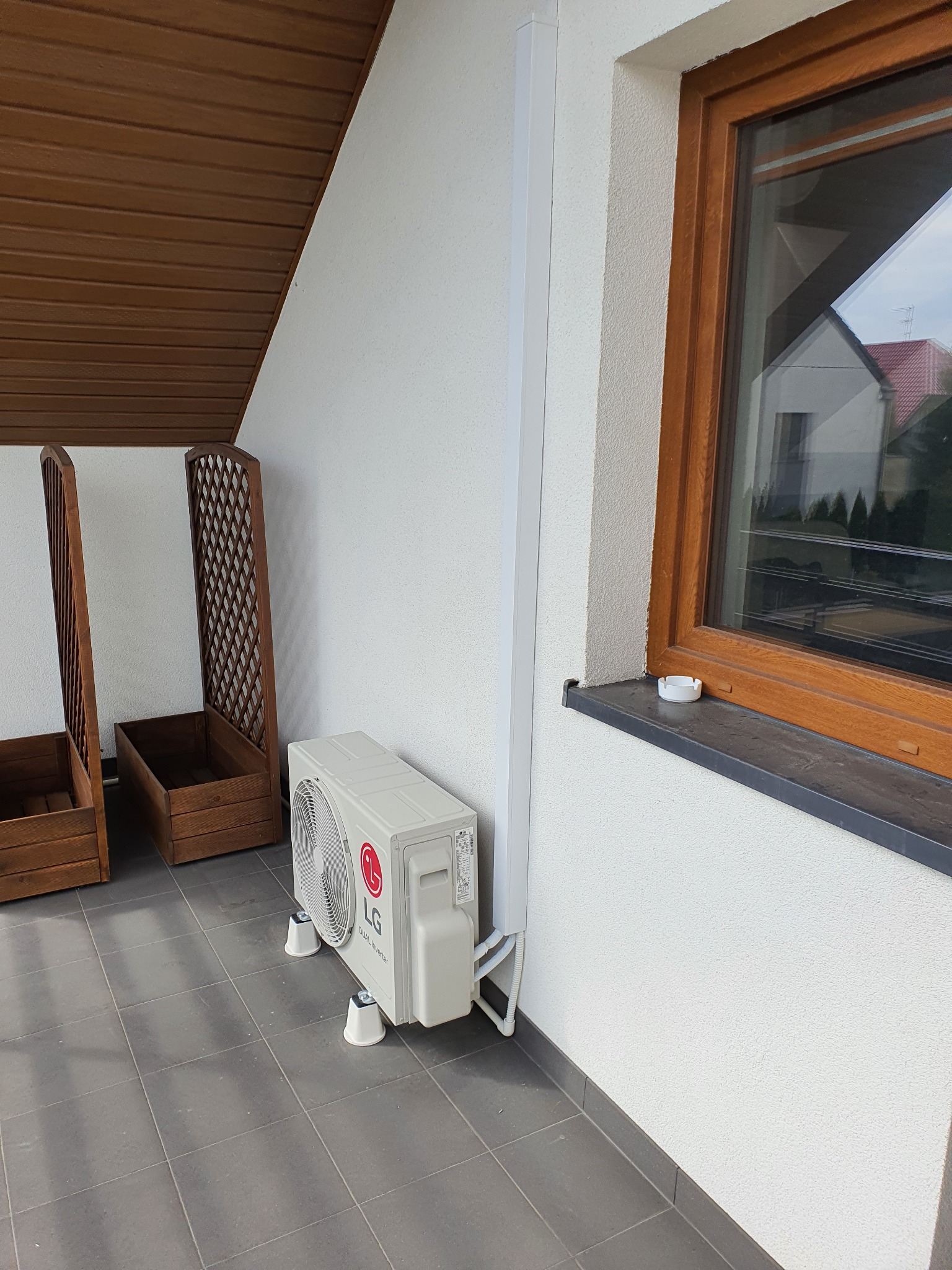 Zewnętrzna jednostka klimatyzacji LG Dual Inverter zamontowana na balkonie, widoczne drewniane skrzynie na rośliny i okno.