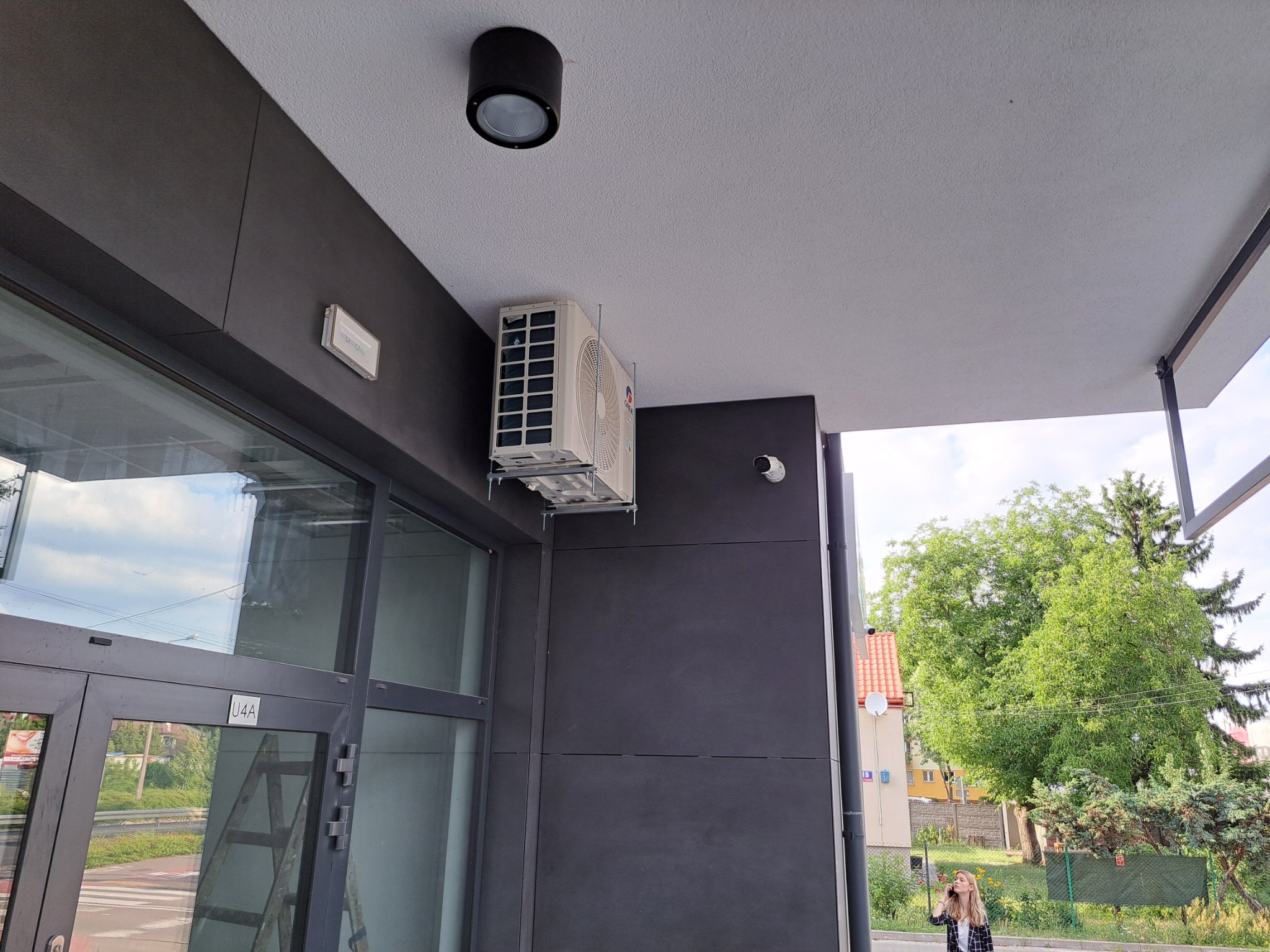 Zewnętrzna jednostka klimatyzatora zamontowana na ścianie budynku komercyjnego, widoczna z dołu, obok okna z numerem U4A, z osobą rozmawiającą przez telefon w tle.