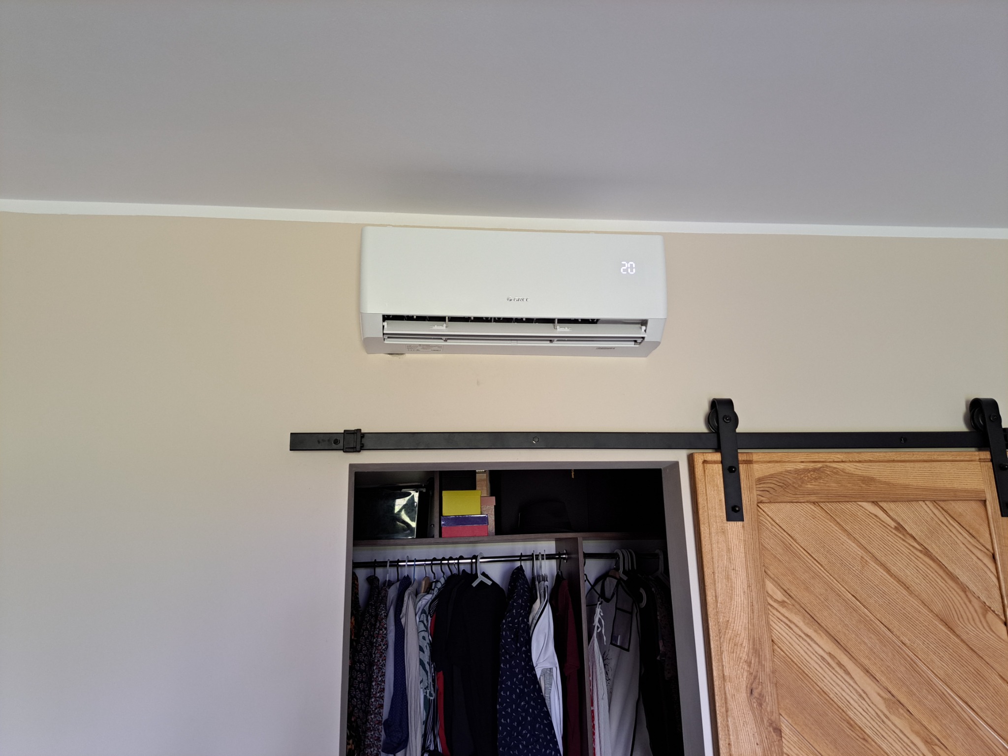 Biały klimatyzator z wyświetlaczem temperatury zamontowany na ścianie nad otwartą szafą z przesuwnymi drzwiami w mieszkaniu w Warszawie.
