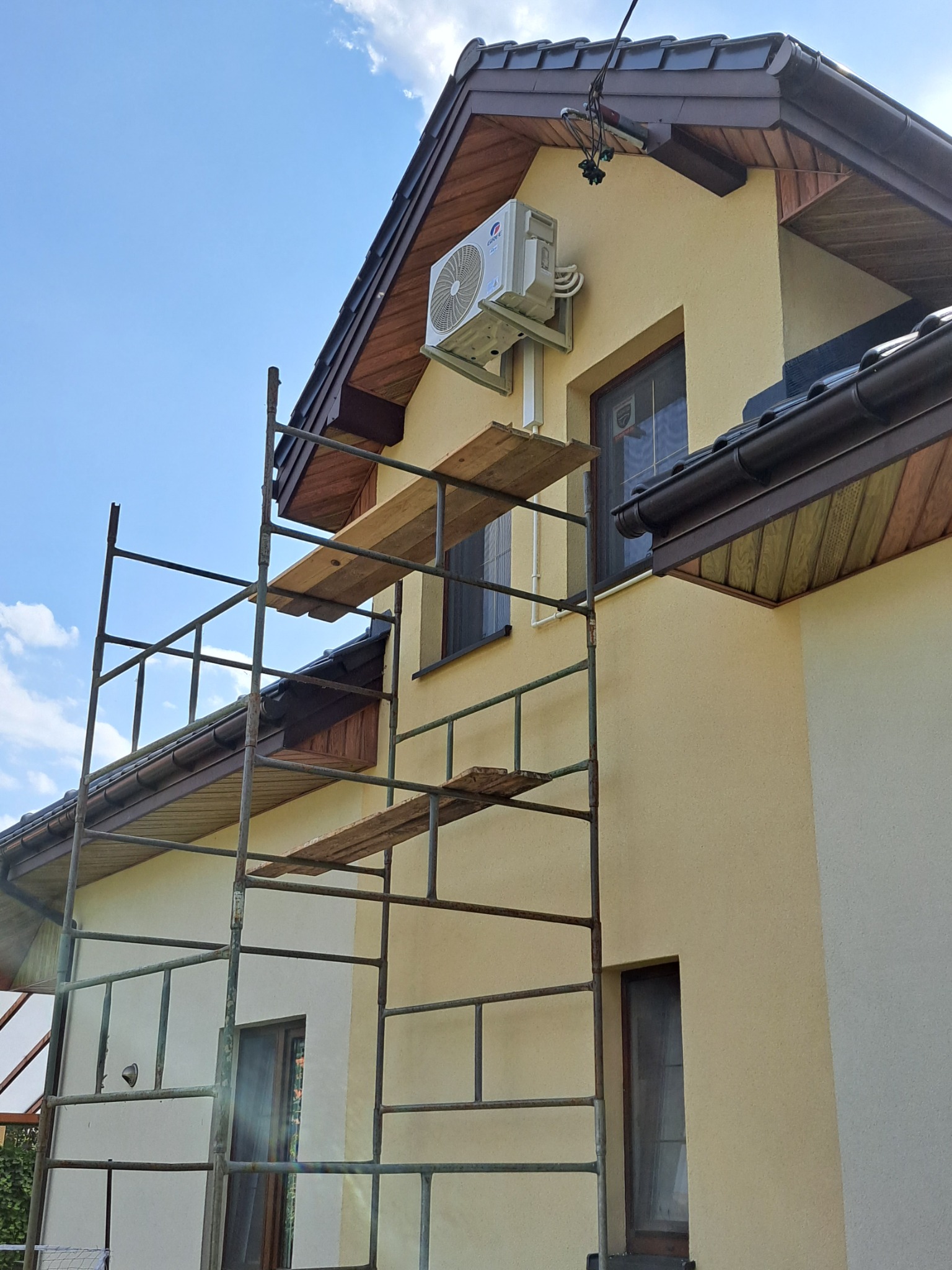 Montaż klimatyzacji na elewacji domu jednorodzinnego, widoczne rusztowanie i jednostka zewnętrzna klimatyzatora marki Gree na ścianie budynku z brązową stolarką okienną.