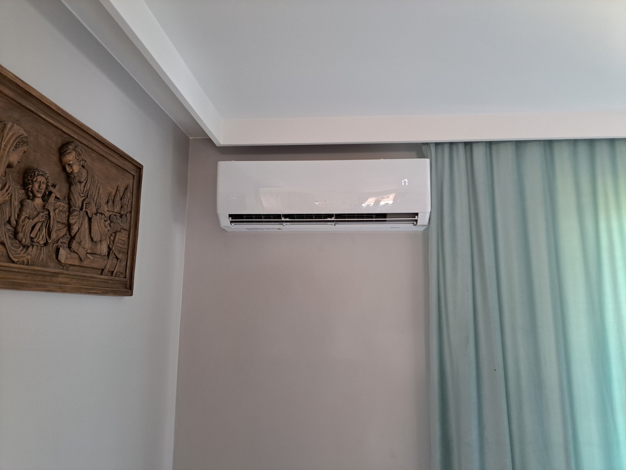 Biały klimatyzator ścienny z wyświetlaczem temperatury 17 stopni Celsjusza, zamontowany w jasnym pomieszczeniu z drewnianą płaskorzeźbą na ścianie i turkusową zasłoną w tle.