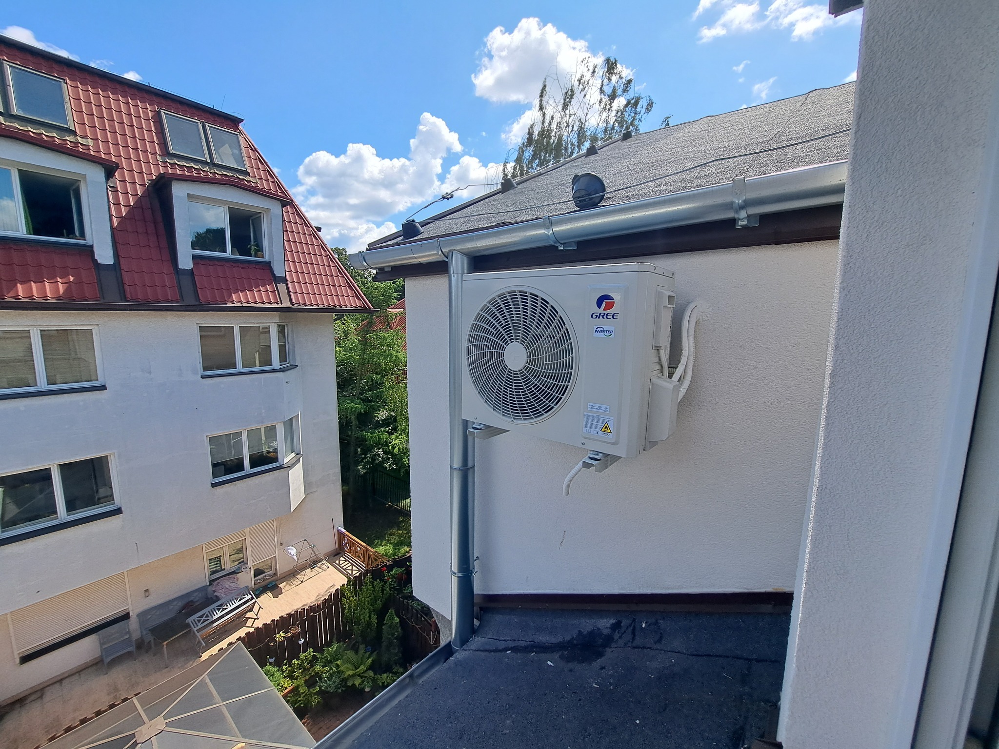 Biały zewnętrzny agregat klimatyzacji marki Gree zamontowany na ścianie budynku, widok z góry na dach, z boku widoczny budynek z czerwonym dachem i okna.