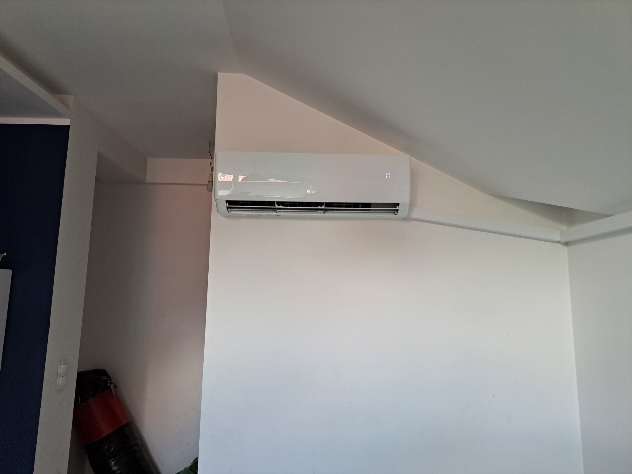 Biały klimatyzator marki Gree zamontowany na białej ścianie pod skosem, widoczny wyświetlacz temperatury ustawionej na 17 stopni, w tle fragment worka bokserskiego.