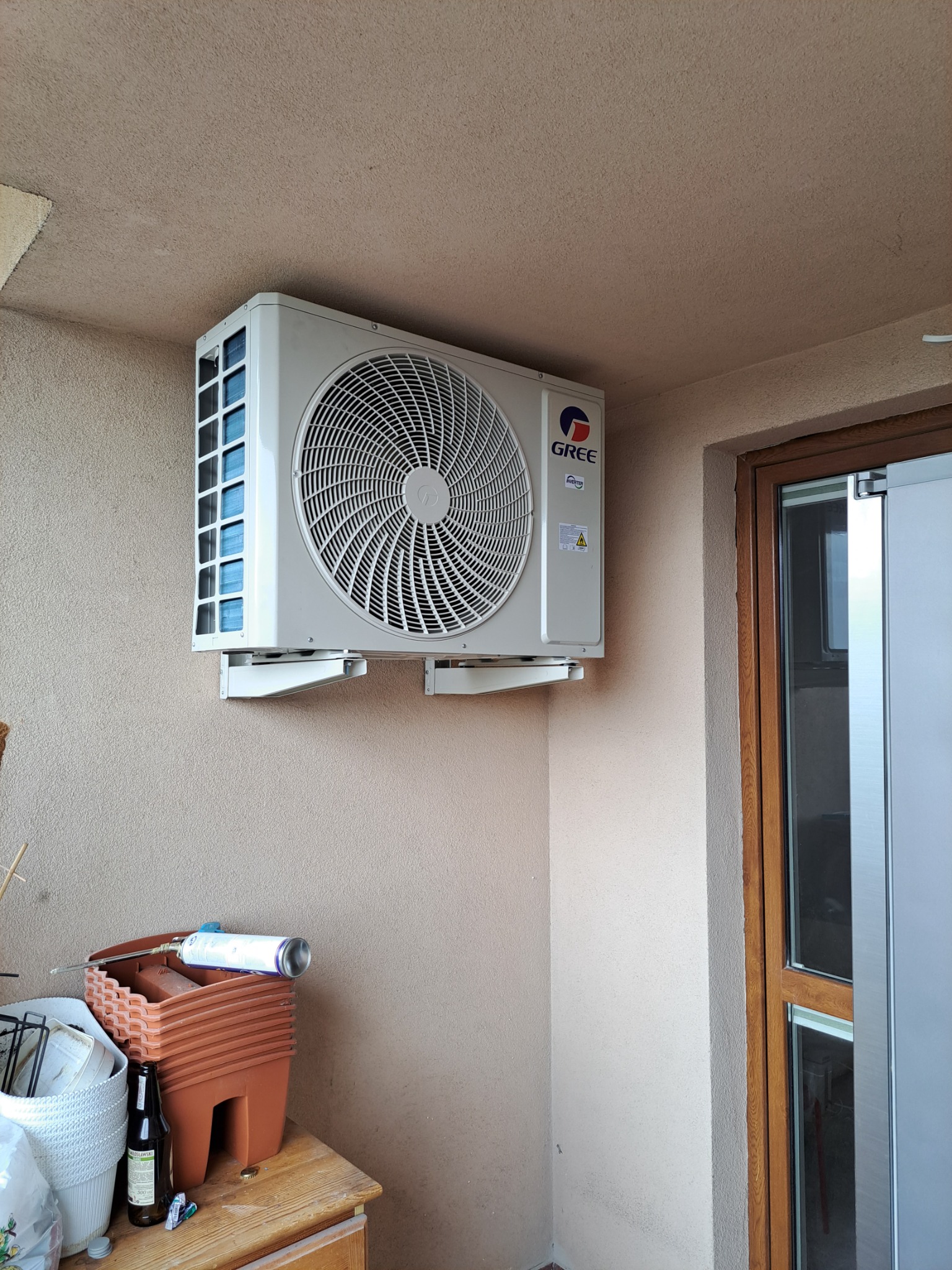 Biała jednostka zewnętrzna klimatyzatora marki Gree zamontowana na beżowej ścianie balkonu, widoczny stolik z doniczkami i butelką.