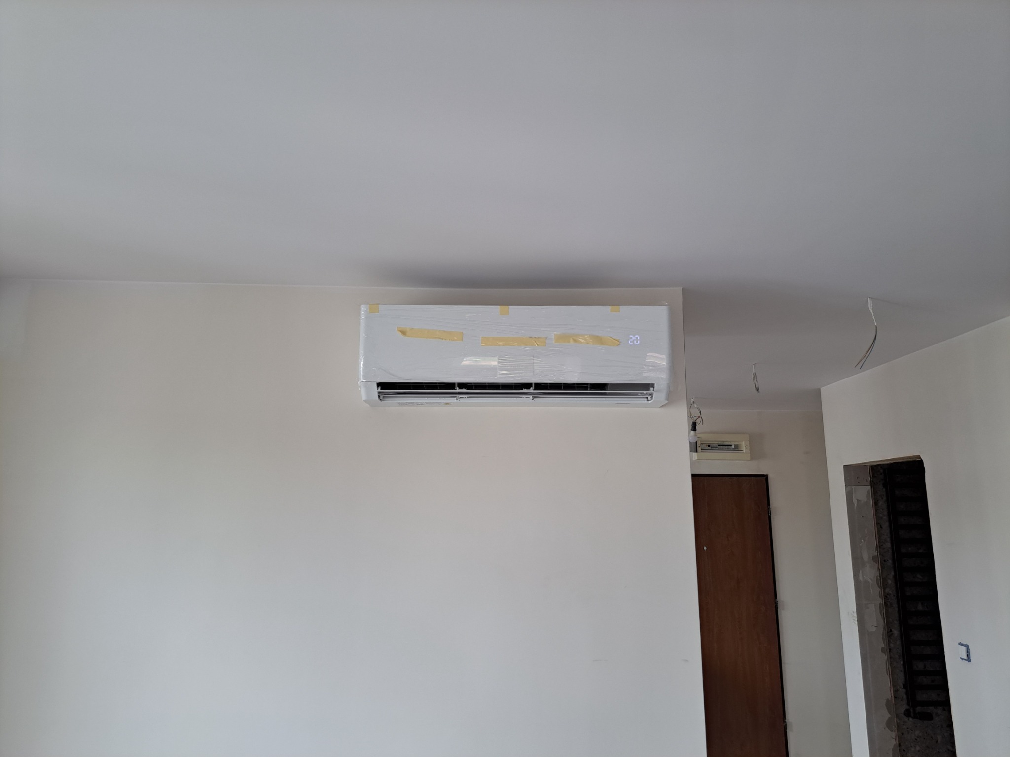 Nowo zamontowana, biała klimatyzacja na ścianie w jasnym pomieszczeniu, zabezpieczona folią i taśmą malarską, widoczny wyświetlacz temperatury.