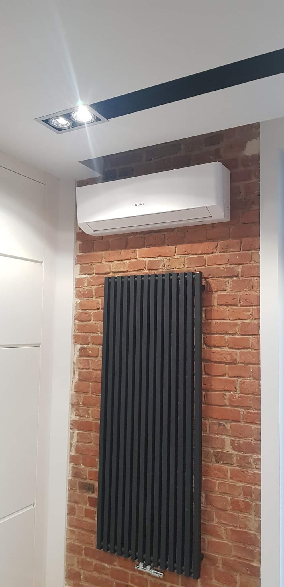 Biała jednostka wewnętrzna klimatyzatora marki Gree zamontowana nad pionowym, czarnym grzejnikiem na tle ściany z czerwonej cegły w nowoczesnym wnętrzu.