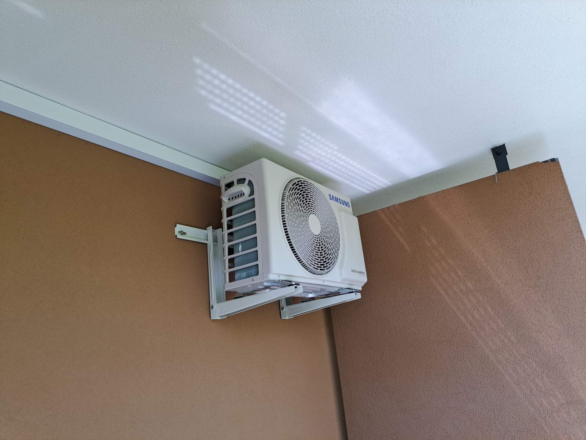 Biała jednostka wewnętrzna klimatyzacji Samsung zamontowana na ścianie w pomieszczeniu z brązowymi ścianami i białym sufitem, widoczne cienie rzucane przez żaluzje.