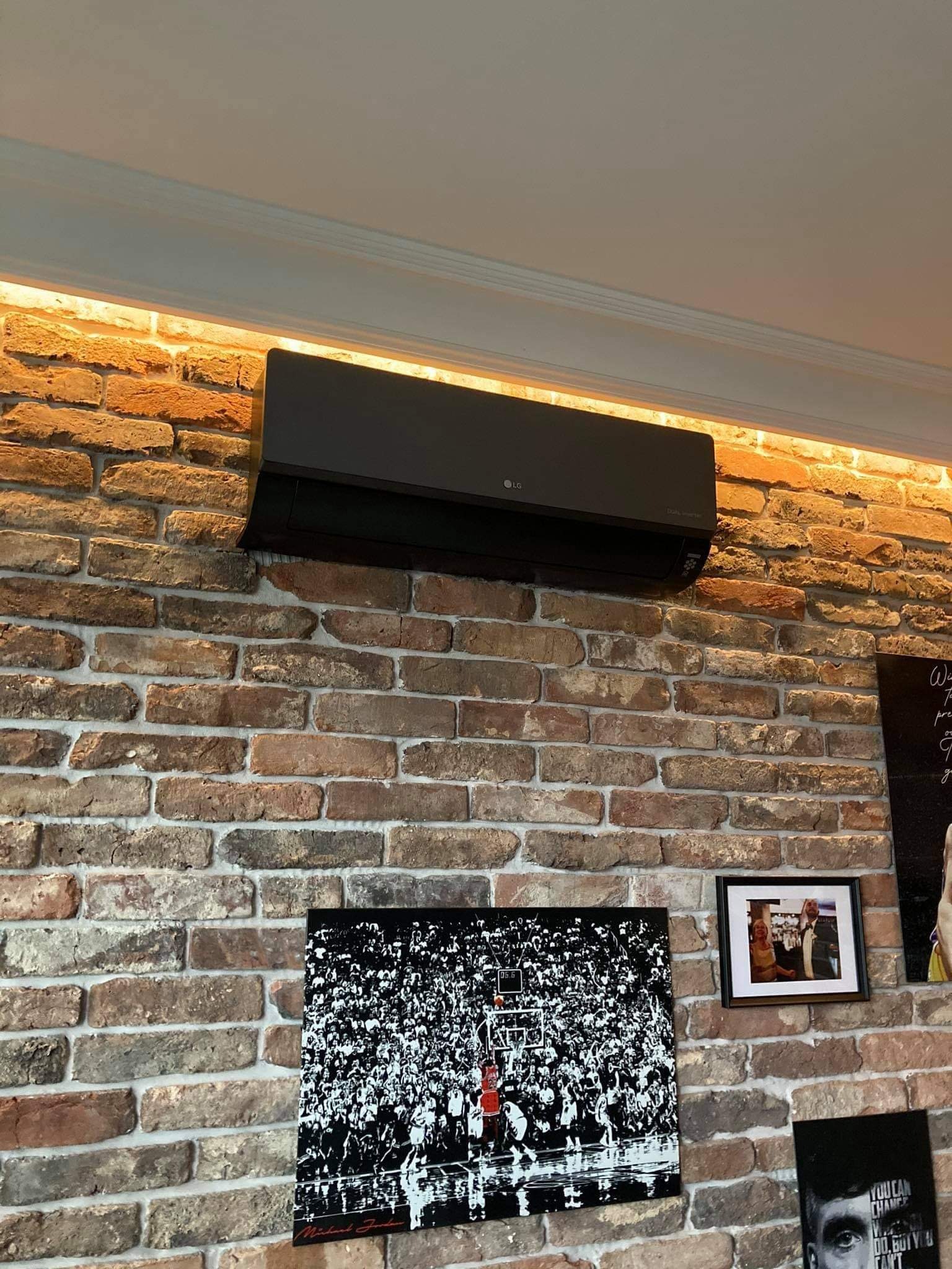 Czarny klimatyzator LG Dual Inverter zamontowany na ceglanej ścianie z oświetleniem LED, pod nim grafiki z Michaelem Jordanem i portret w ramce.