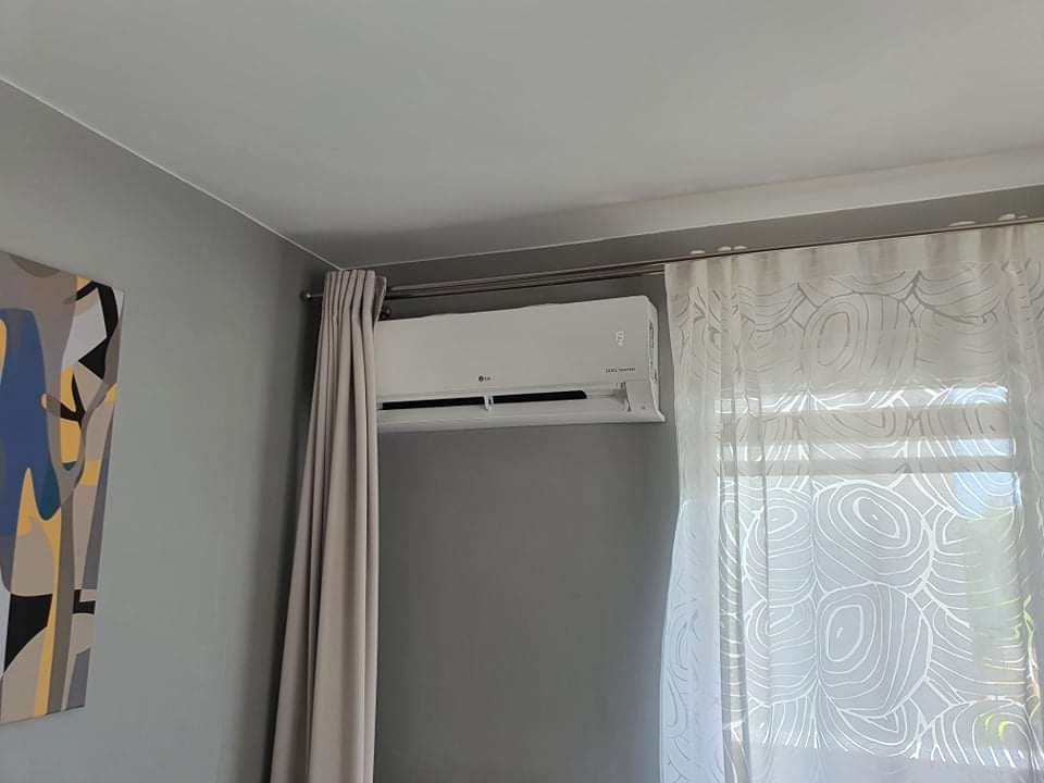 Zamontowany klimatyzator LG na szarej ścianie w pokoju, obok okna z białymi, wzorzystymi zasłonami i szarą zasłoną, z abstrakcyjnym obrazem wiszącym po lewej stronie.