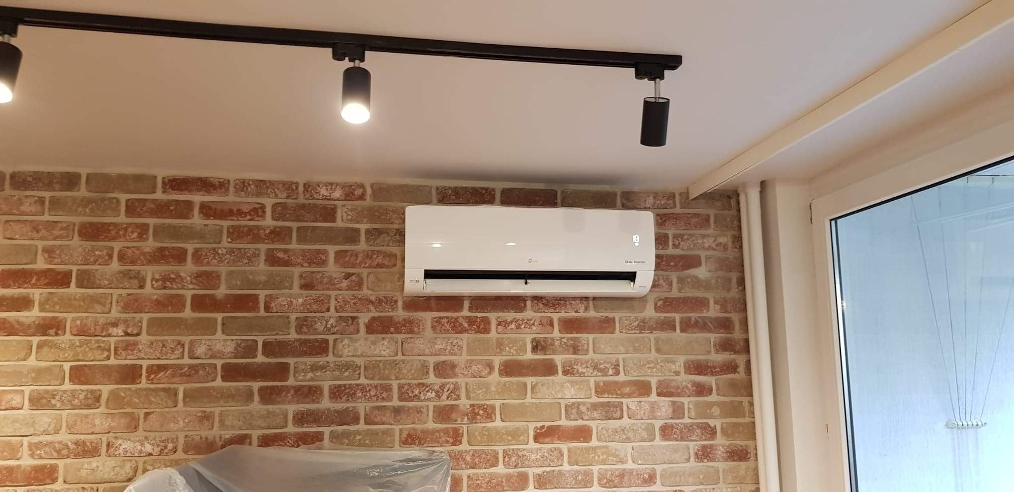 Biały klimatyzator LG zamontowany na ścianie z cegły dekoracyjnej, oświetlony reflektorami szynowymi.