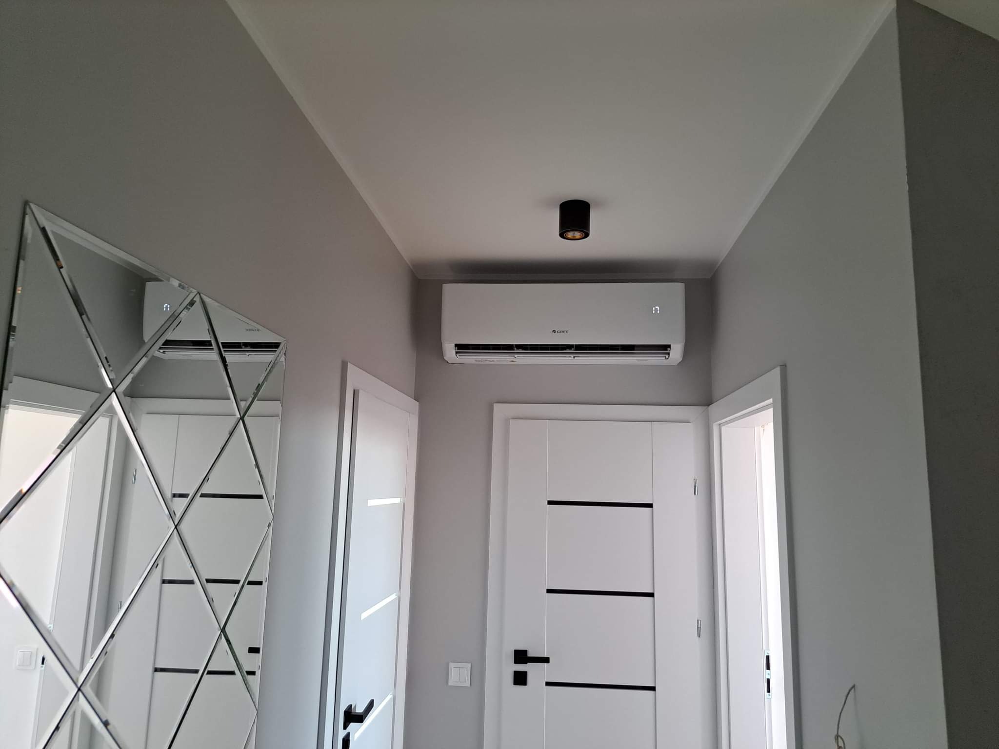 Biała jednostka klimatyzacyjna Gree zamontowana nad białą, nowoczesną drzwiami w szarym pomieszczeniu, odbicie w lustrze diamentowym.