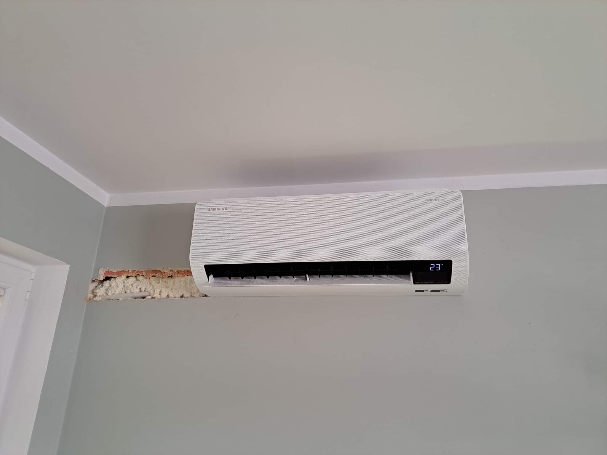 Biała jednostka klimatyzacji Samsung zamontowana na szarej ścianie, z widocznym fragmentem ściany wypełnionym pianką montażową po instalacji.
