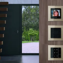 Panel sterowania z wideodomofonem, głośnikiem i przyciskami funkcyjnymi zamontowany na drewnianej ścianie w nowoczesnym wnętrzu z widokiem na zewnątrz przez otwarte drzwi.