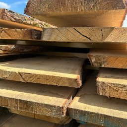 Centrum Drzewne Drewienko - Stos desek z widocznymi słojami drewna i korą, ułożonych warstwami pod błękitnym niebem. Naturalna tekstura drewna w ciepłym oświetleniu.