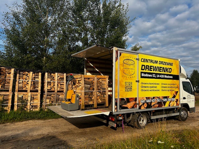 Dostawa drewna kominkowego: samochód dostawczy z logo firmy 'Drewienko' rozładowuje palety drewna za pomocą wózka paletowego. W tle drzewa.