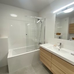 B&K GROUP SP. Z O.O - Nowoczesna łazienka z białymi płytkami, szklanym panelem prysznicowym nad wanną i drewnianą szafką pod umywalką, oświetlona światłem LED.