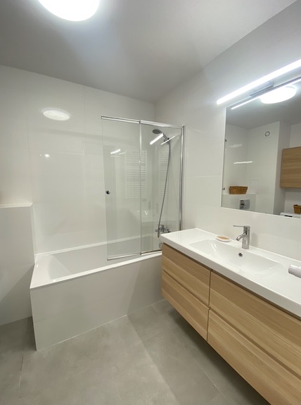 Nowoczesna łazienka z białymi płytkami, szklanym panelem prysznicowym nad wanną i drewnianą szafką pod umywalką, oświetlona światłem LED.