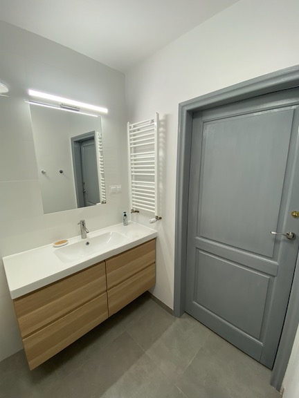 Nowoczesna łazienka z drewnianą szafką pod umywalką, grzejnikiem drabinkowym i szarą, panelową ścianą.