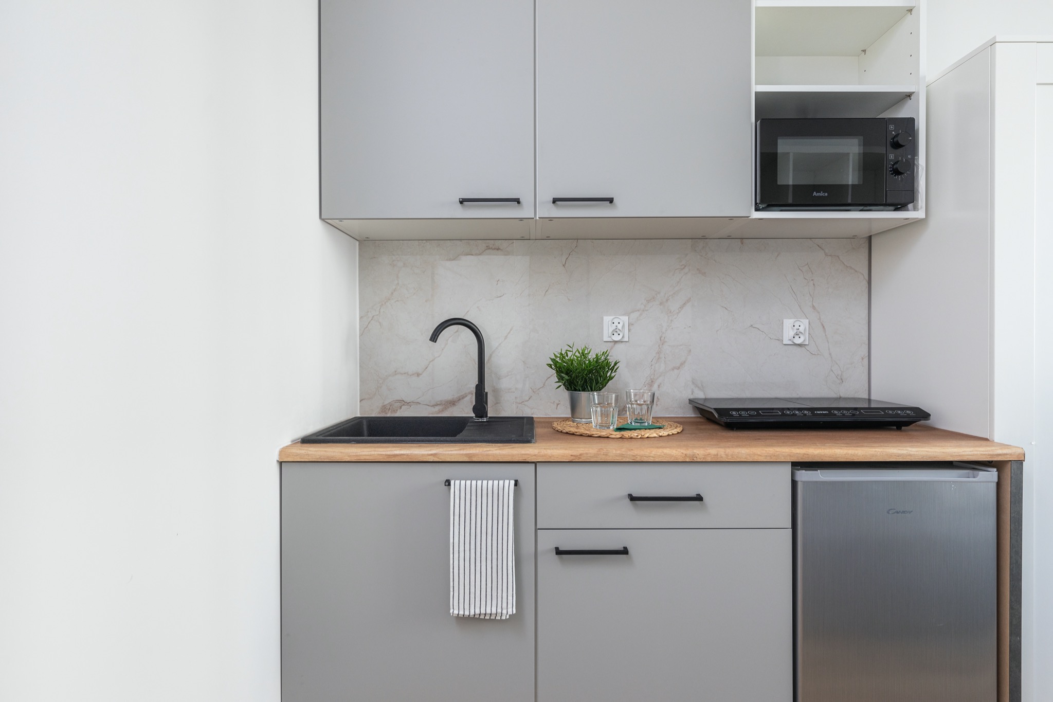 Szara, minimalistyczna kuchnia z czarnym zlewem i baterią, drewnianym blatem, płytą indukcyjną, mikrofalówką Amica i małą lodówką Candy, uzupełniona zieloną rośliną w doniczce i ręcznikiem w paski.