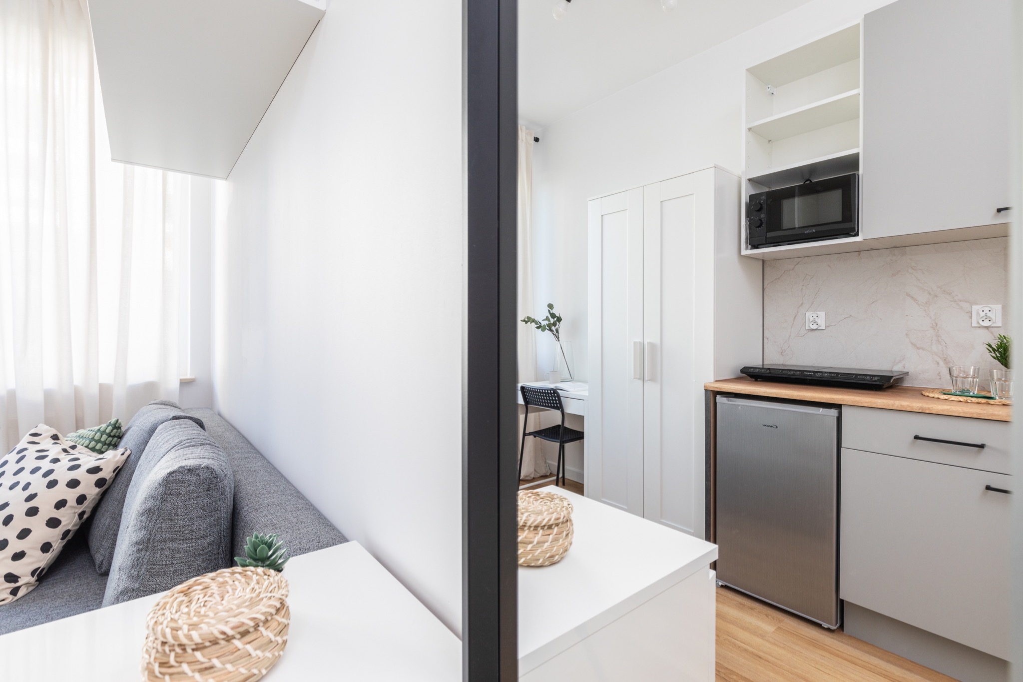 Nowoczesne studio: szara sofa z poduszkami w kropki, białe ściany, aneks kuchenny z szarymi szafkami, mikrofalówką i małą lodówką, przedzielone czarną ramą.