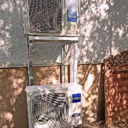 Dwa klimatyzatory Haier zamontowane jeden nad drugim na metalowej konstrukcji, cień drzewa na elewacji, widok na drewniany płot.