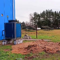 Zewnętrzna jednostka pompy ciepła zamontowana na betonowym fundamencie przy błękitnej ścianie budynku, panele słoneczne w tle, teren po robotach ziemnych.