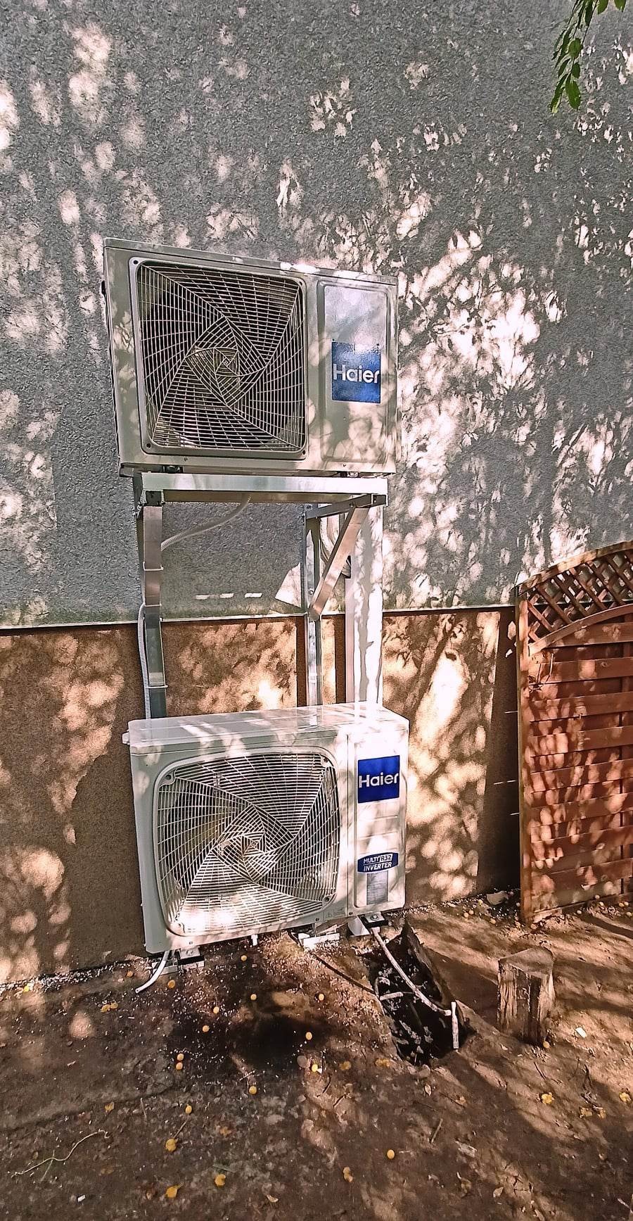 Dwa klimatyzatory Haier zamontowane jeden nad drugim na metalowej konstrukcji, cień drzewa na elewacji, widok na drewniany płot.