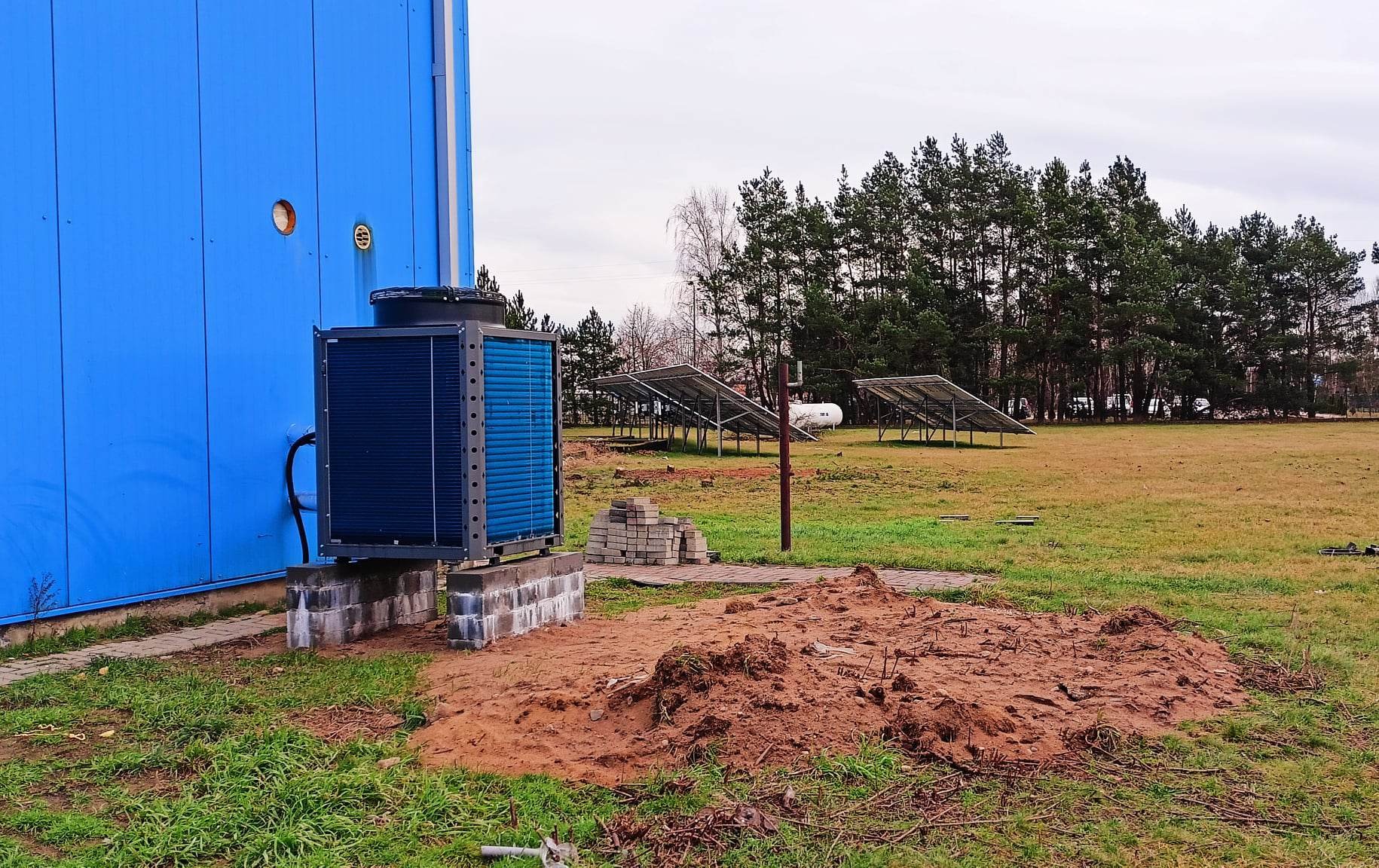 Zewnętrzna jednostka pompy ciepła zamontowana na betonowym fundamencie przy błękitnej ścianie budynku, panele słoneczne w tle, teren po robotach ziemnych.
