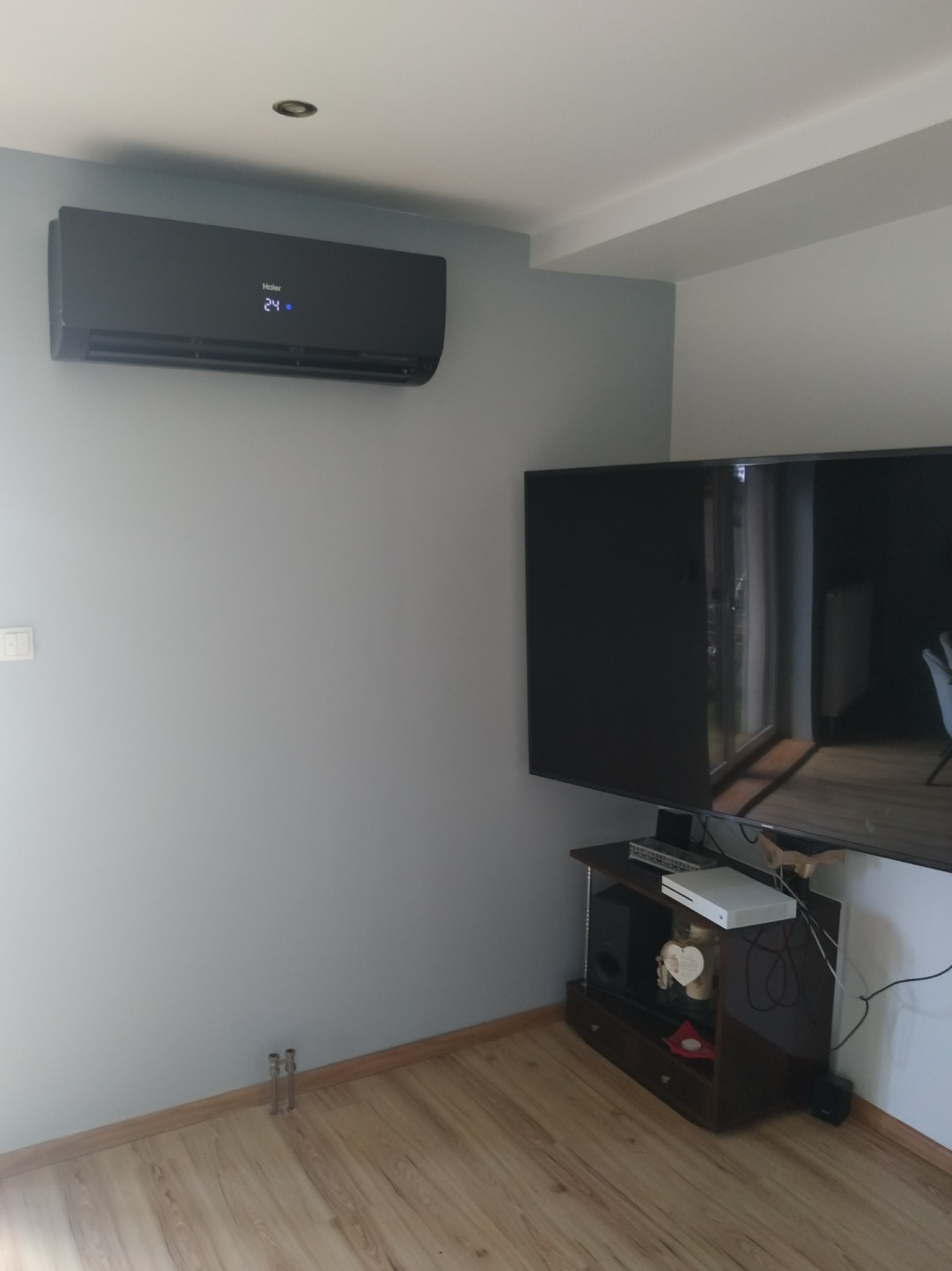 Czarny klimatyzator Haier zamontowany na szarej ścianie w pokoju, widoczny wyświetlacz z temperaturą 24 stopnie, obok telewizor na stoliku RTV.