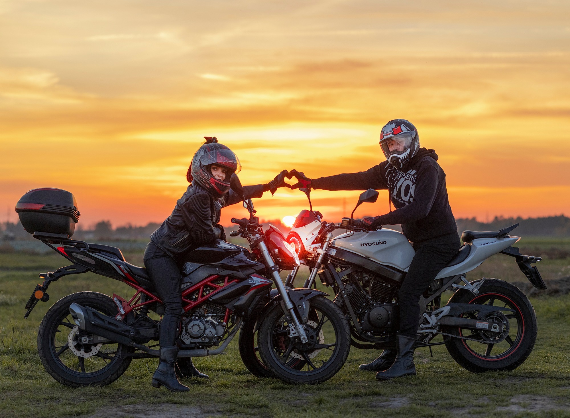 Para w kaskach motocyklowych siedzi na motocyklach na tle zachodzącego słońca, tworząc serce dłońmi nad przednimi światłami.