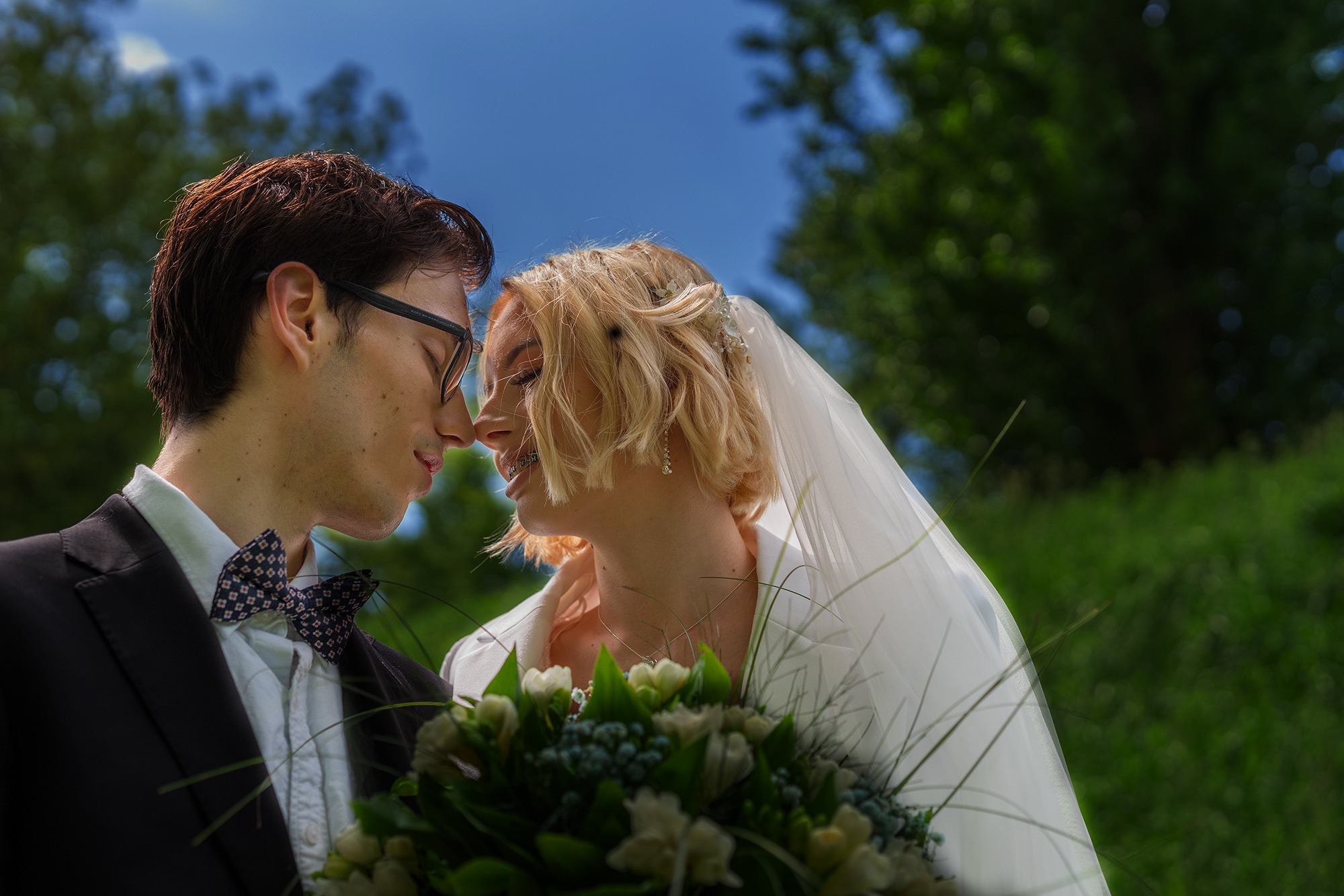 Młoda para w zbliżeniu, panna młoda z blond włosami i welonem, pan młody w okularach i muszce, delikatny bukiet ślubny, tło z zieleni i błękitnego nieba.