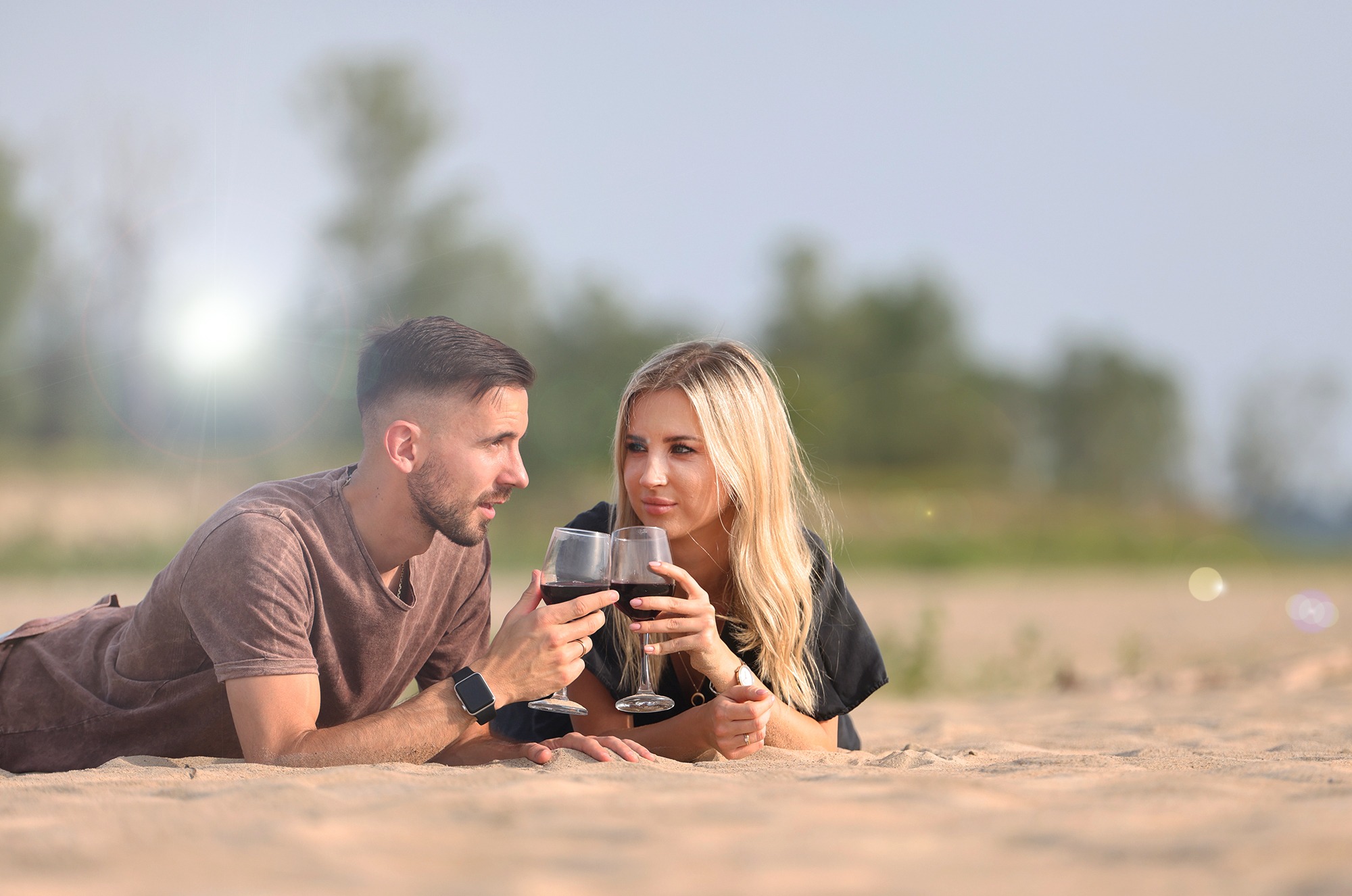 Para leżąca na piasku, trzymająca kieliszki z czerwonym winem, spoglądająca na siebie z uśmiechem, w tle rozmyty krajobraz.