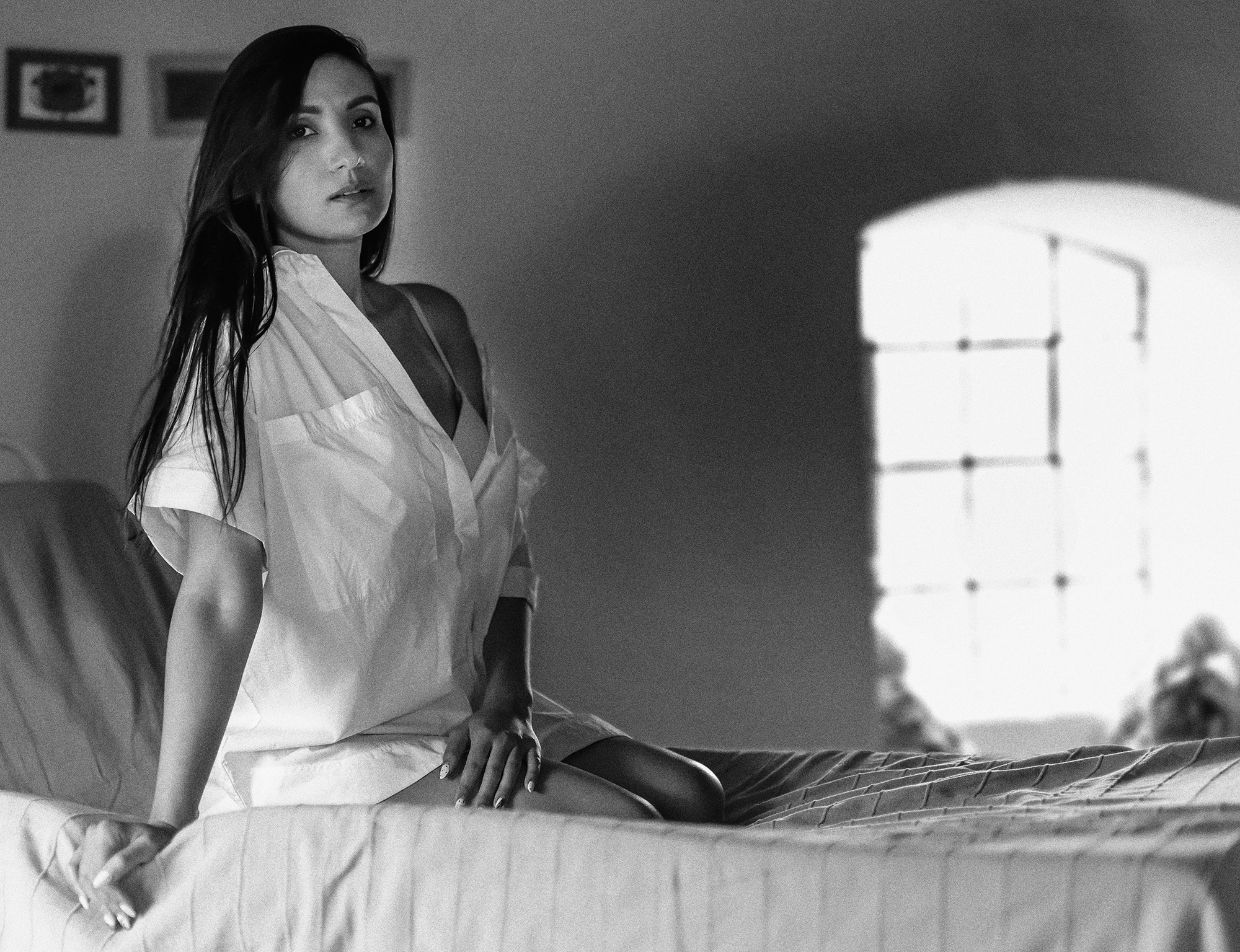Czarno-białe zdjęcie kobiety siedzącej na łóżku w białej koszuli, z rozpuszczonymi włosami, spoglądającej w obiektyw, z oknem w tle.