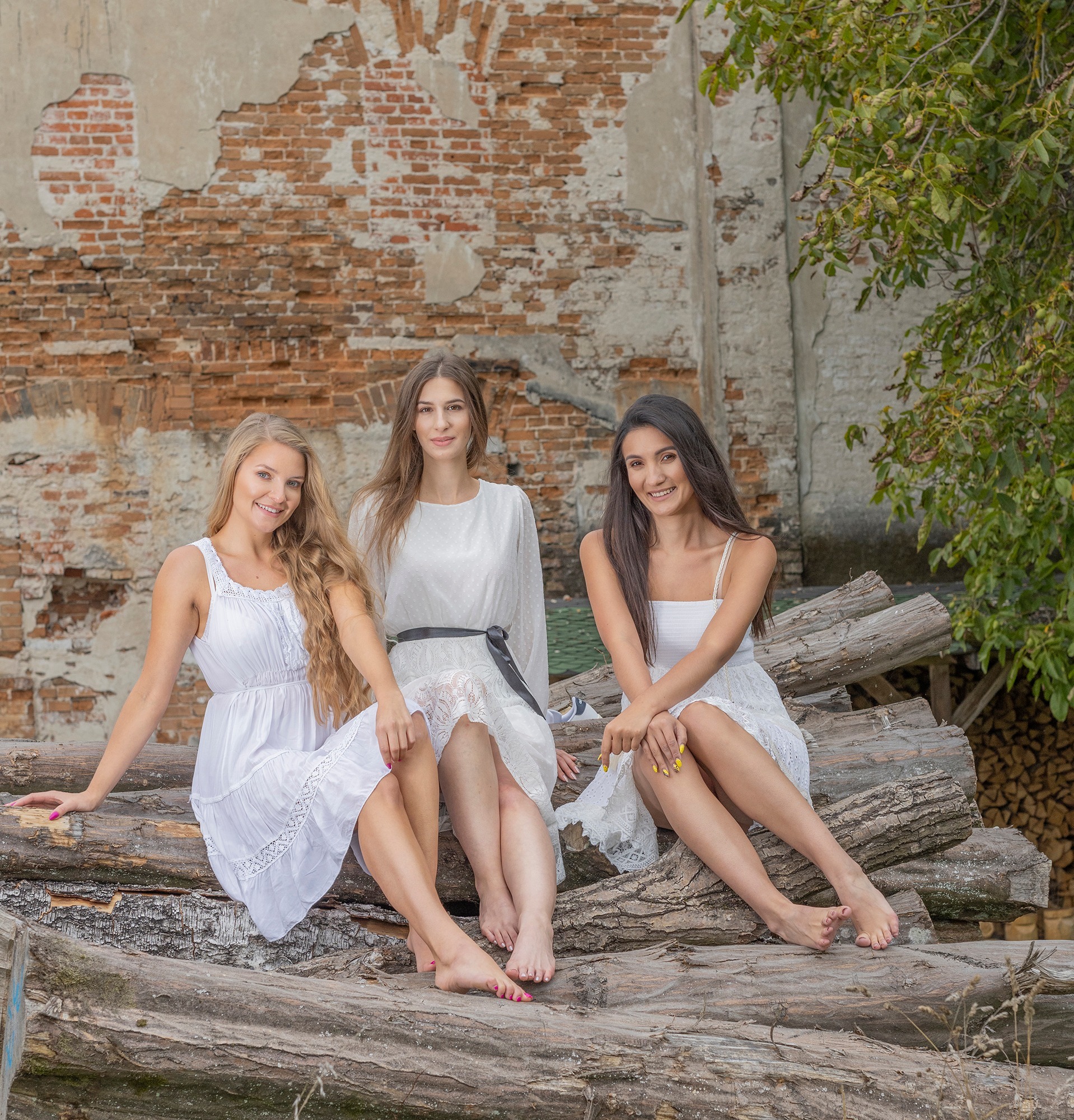 Trzy uśmiechnięte kobiety w białych sukienkach pozują siedząc na stosie drewnianych bali na tle starej, ceglanej ściany i drzewa z zielonymi liśćmi.