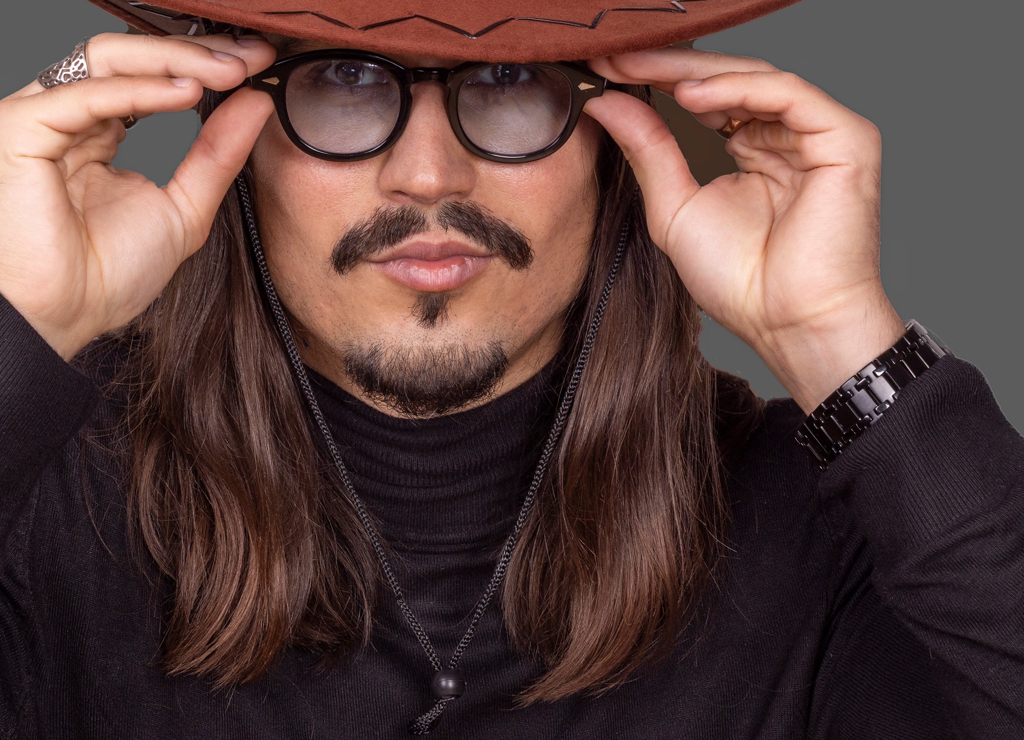 Portret mężczyzny z długimi włosami, wąsami i bródką, w brązowym kapeluszu i okularach, dotykającego oprawek okularów palcami, na szarym tle.