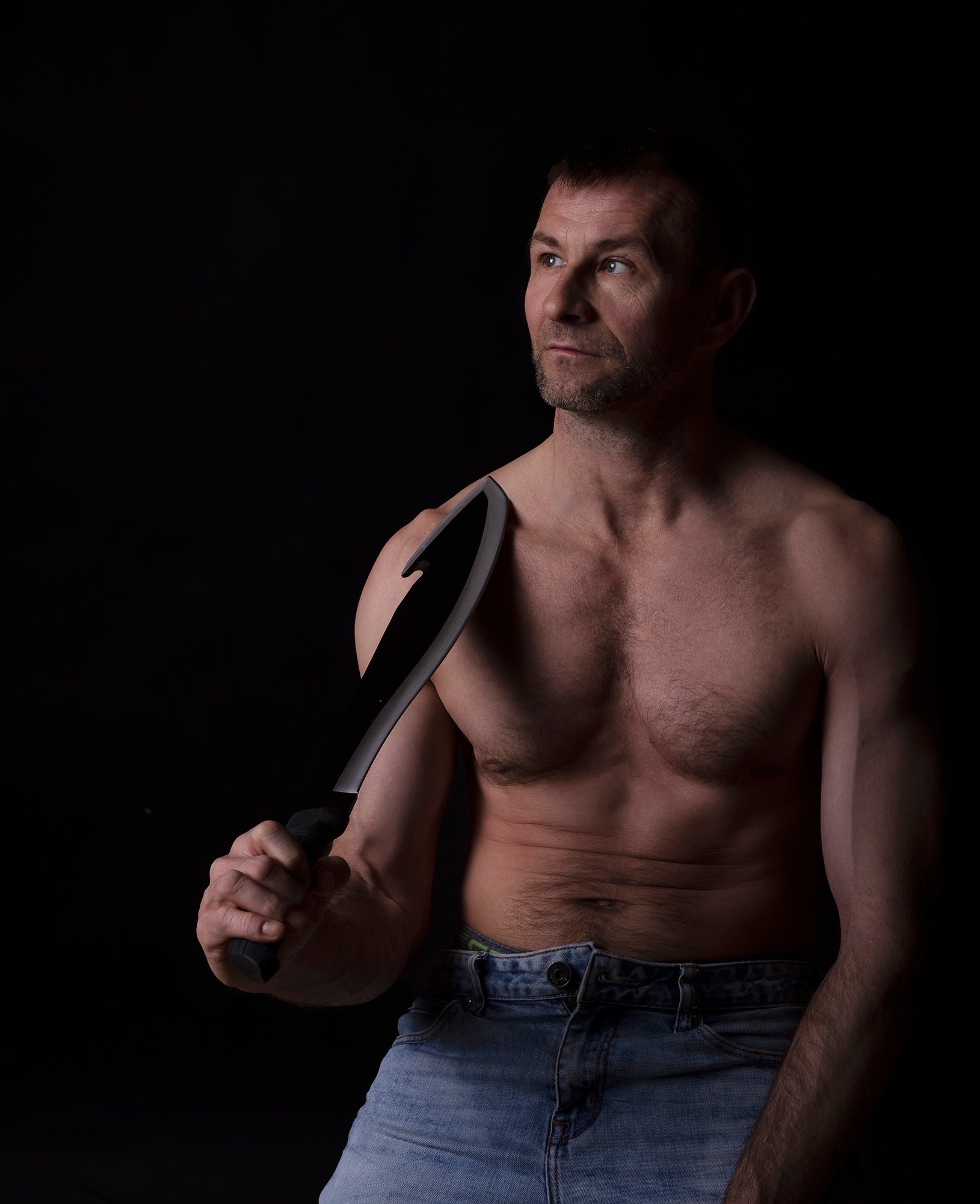 Mężczyzna bez koszuli, w dżinsach, trzymający duży nóż o zakrzywionym ostrzu, spoglądający w górę w ciemnym otoczeniu.