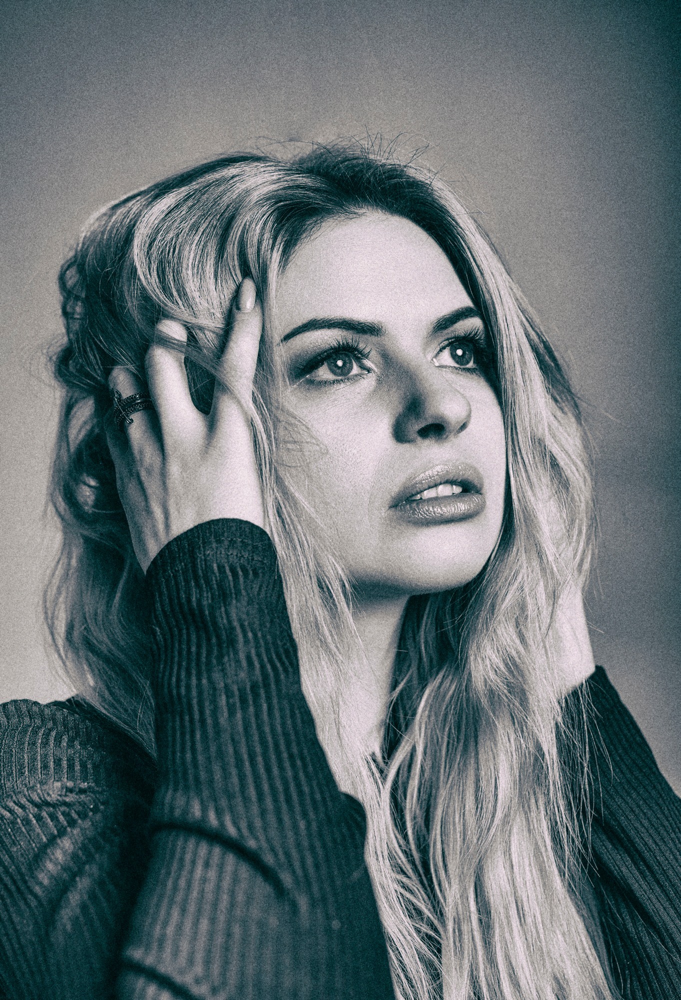 Portret kobiety z blond włosami, spoglądającej w górę, z dłonią we włosach i pierścionkiem na palcu, w czarno-białej tonacji.