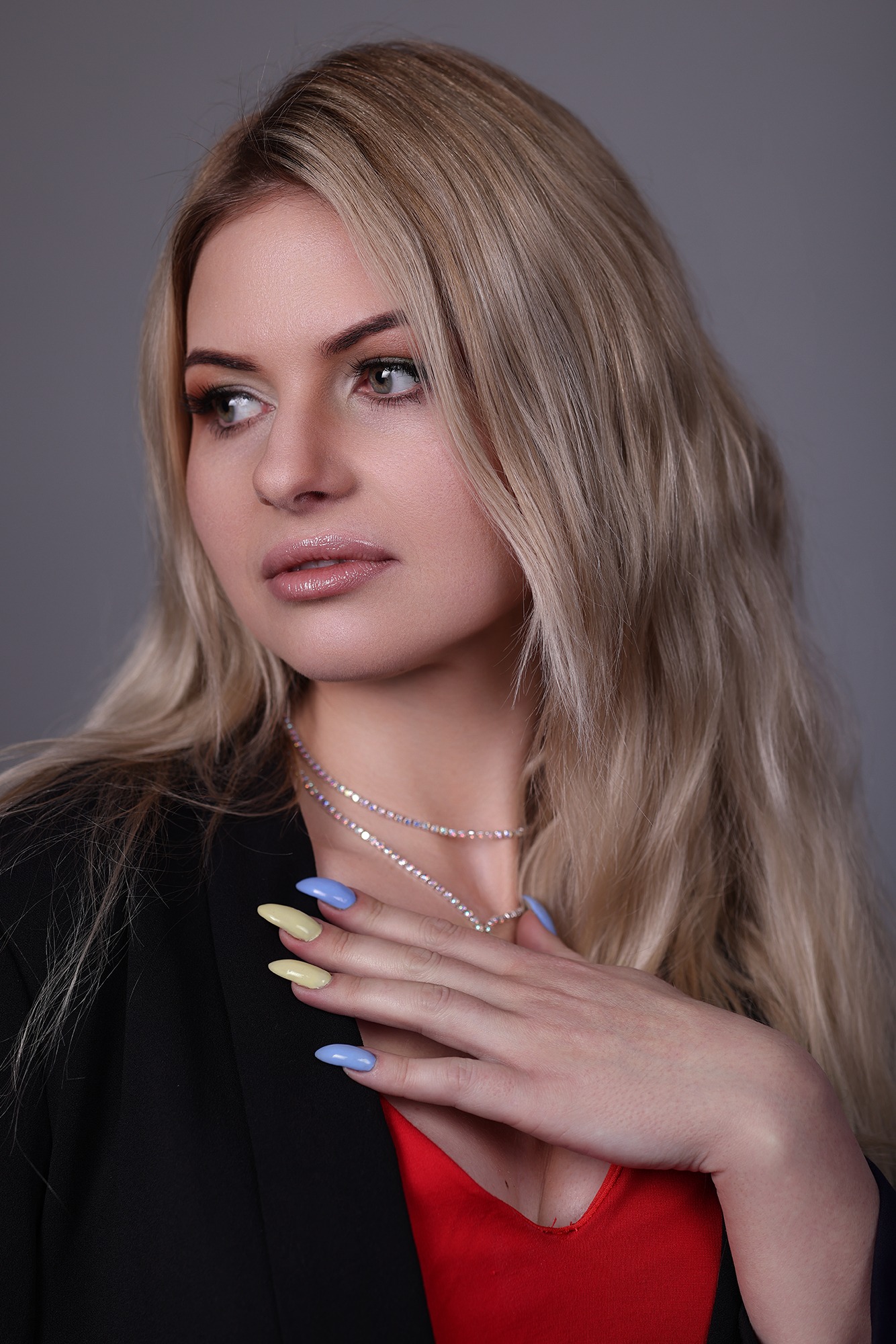 Portret kobiety z blond włosami, w czarnej marynarce i z delikatnym makijażem, z ręką na szyi, eksponujący biżuterię i manicure w pastelowych kolorach.