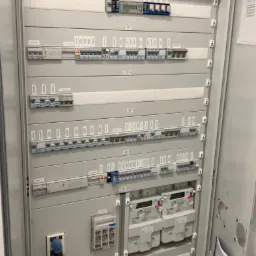 Otwarta szafa rozdzielcza z zainstalowanymi licznikami energii elektrycznej, bezpiecznikami i wyłącznikami różnicowoprądowymi, widoczne przewody w korytkach na górze.