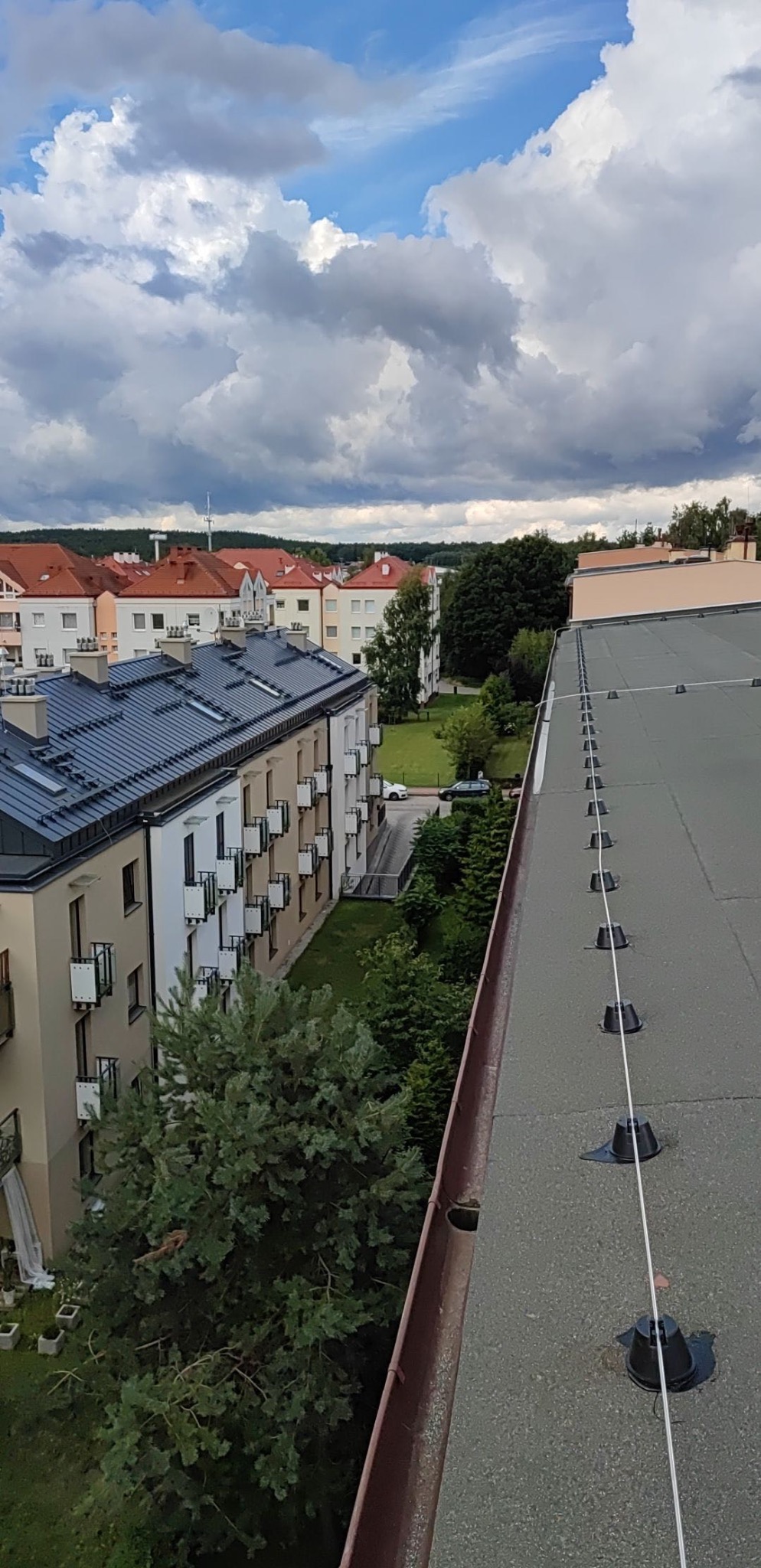 Widok z góry na płaski dach z zamontowaną instalacją odgromową, w tle budynki mieszkalne i pochmurne niebo.