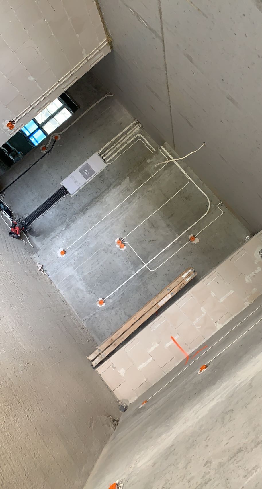 Surowy stan wnętrza z widoczną instalacją elektryczną: kable w białych peszlach prowadzone po suficie i ścianach, pomarańczowe puszki pod gniazdka i włączniki, skrzynka rozdzielcza na ścianie...