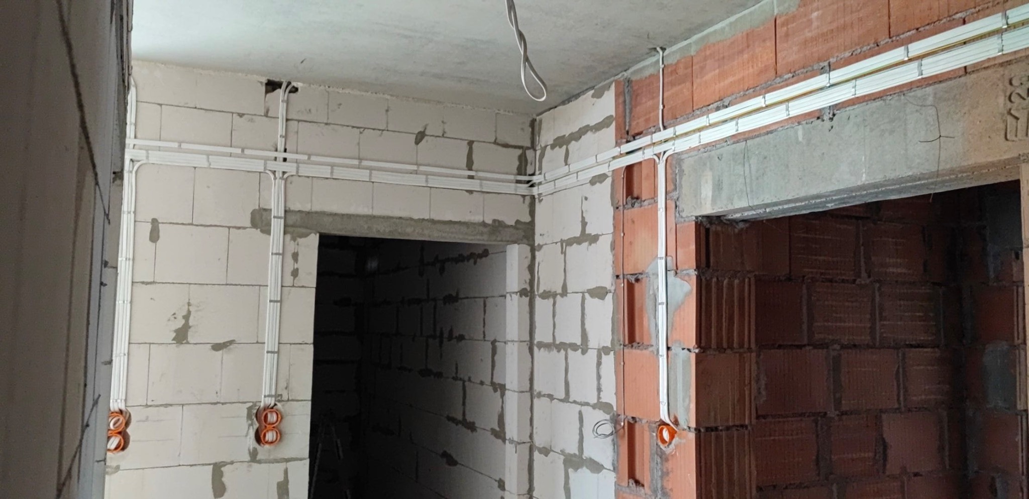 Wnętrze w trakcie remontu z widocznymi białymi listwami elektroinstalacyjnymi ułożonymi na ścianach z białego i czerwonego pustaka, prowadzącymi do puszek instalacyjnych w kolorze pomarańczowym.