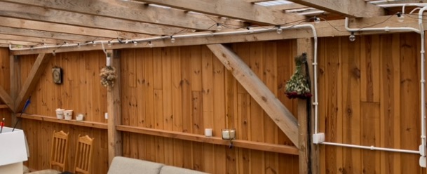 Drewniana pergola z oświetleniem zewnętrznym: białe przewody elektryczne prowadzone po drewnianych elementach konstrukcyjnych, żarówki dekoracyjne, drewniana ściana w tle.