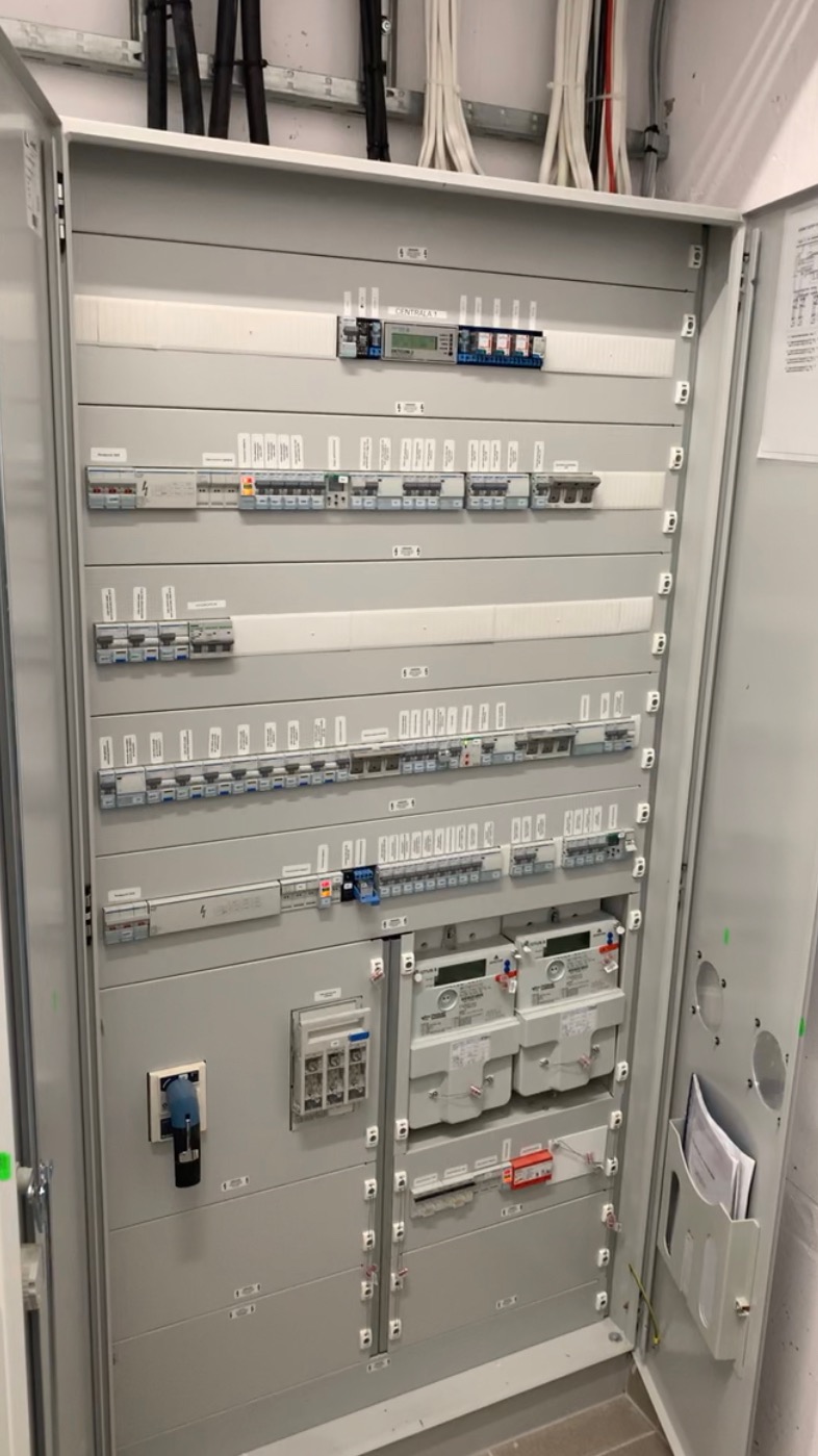Otwarta szafa rozdzielcza z zainstalowanymi licznikami energii elektrycznej, bezpiecznikami i wyłącznikami różnicowoprądowymi, widoczne przewody w korytkach na górze.
