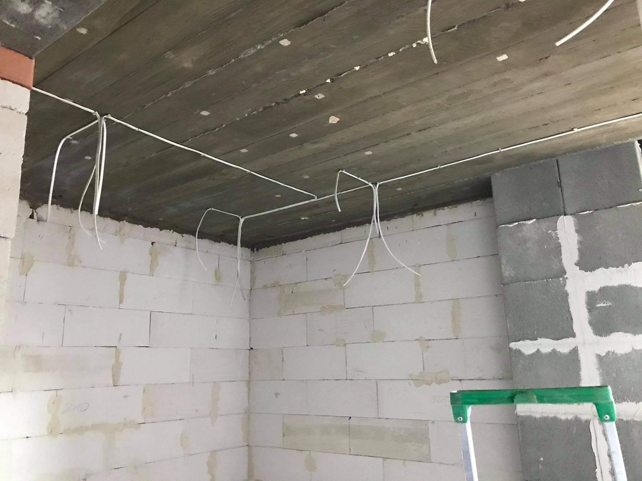 Surowy betonowy strop z widocznymi przewodami elektrycznymi w białej osłonie, ściany z białych bloczków betonowych, częściowo otynkowane, zielona drabina oparta o ścianę w rogu pomieszczenia.