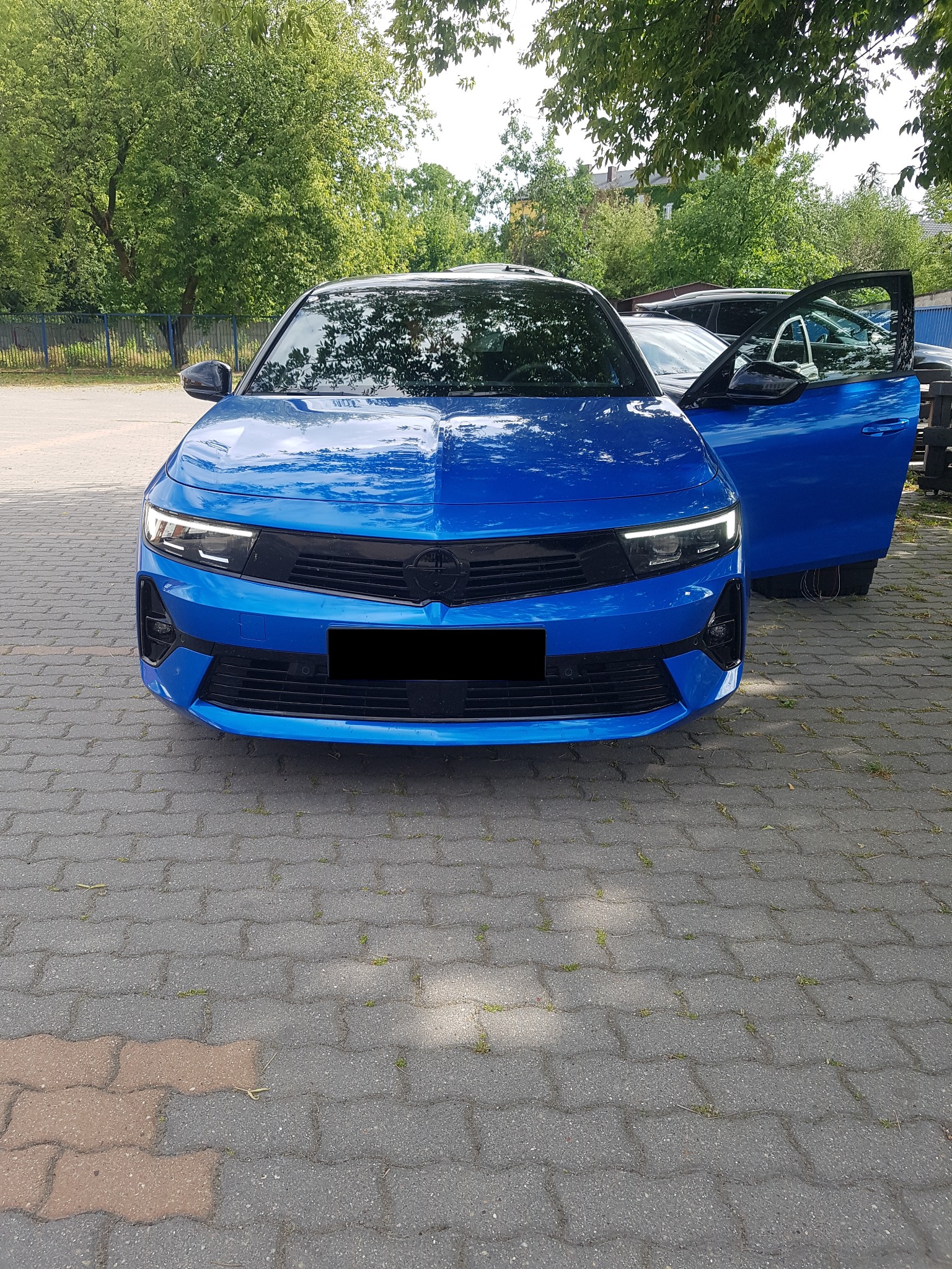 Niebieski samochód osobowy z otwartymi drzwiami po stronie pasażera, zaparkowany na brukowanym placu, widok z przodu.